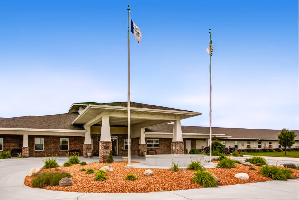 Exterior view of Addington Place of Carroll, showing a single-story brick building with a covered entrance supported by white pillars. Two flagpoles with flags stand in front of the entrance, surrounded by landscaped mulch beds with green plants and rocks. The sky is clear and blue.