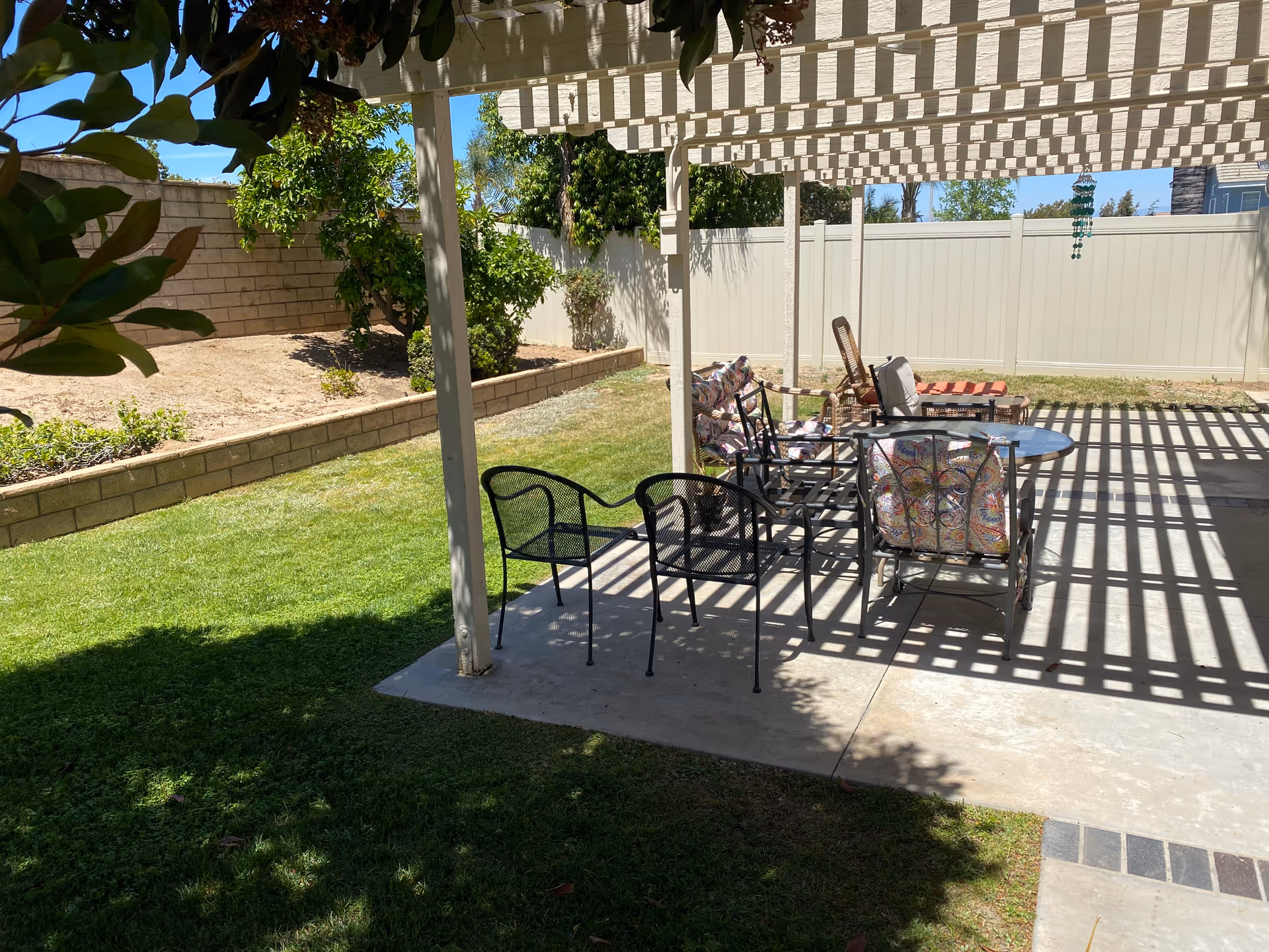 Outdoor patio area with a white pergola casting striped shadows on the concrete floor. Several chairs, including metal and cushioned ones, are arranged around a small round table. The patio is adjacent to a grassy lawn and bordered by a low brick wall and a white fence. Trees and plants are visible in the background under a clear blue sky.