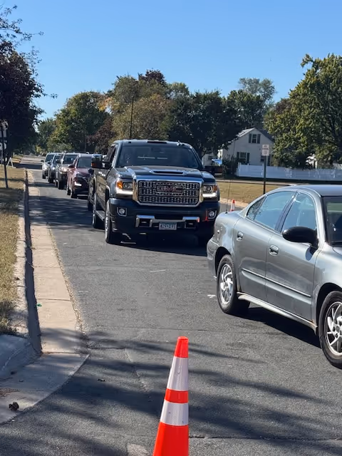 Line of vehicles parked along a residential street with a traffic cone in foreground.