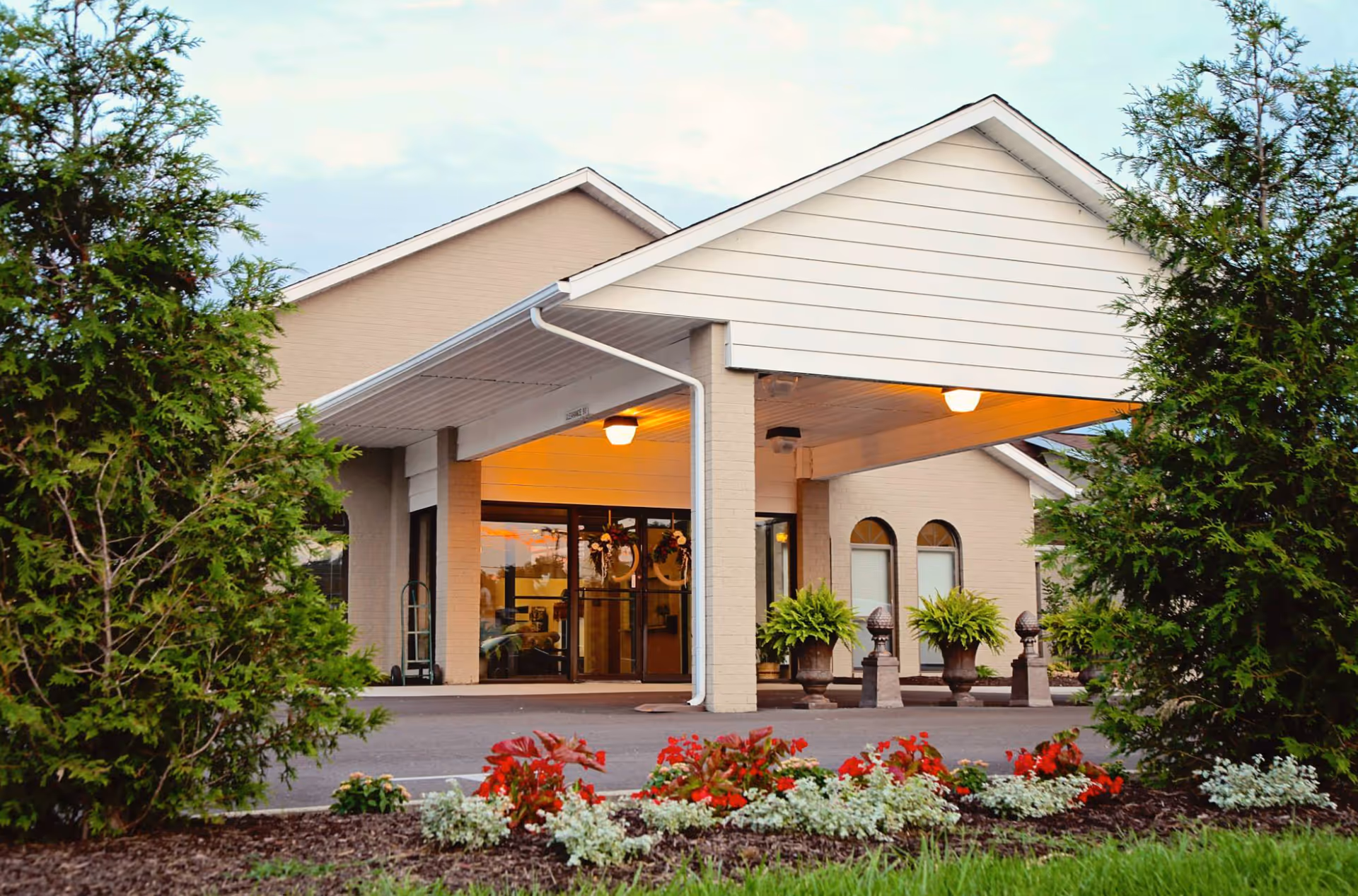 Entrance of Commonwealth Senior Living at Radford showing a covered drop-off area with glass doors, potted plants, and decorative greenery. The building exterior is light-colored with a peaked roof, surrounded by trees and a flower bed with red and white flowers in the foreground.