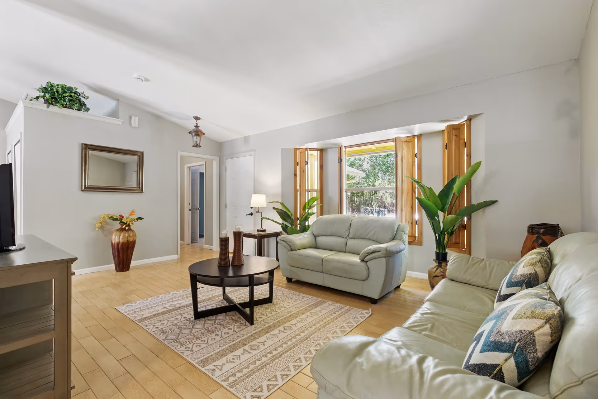 Bright living room with light-colored sofas, coffee table, plants, and large window with shutters.