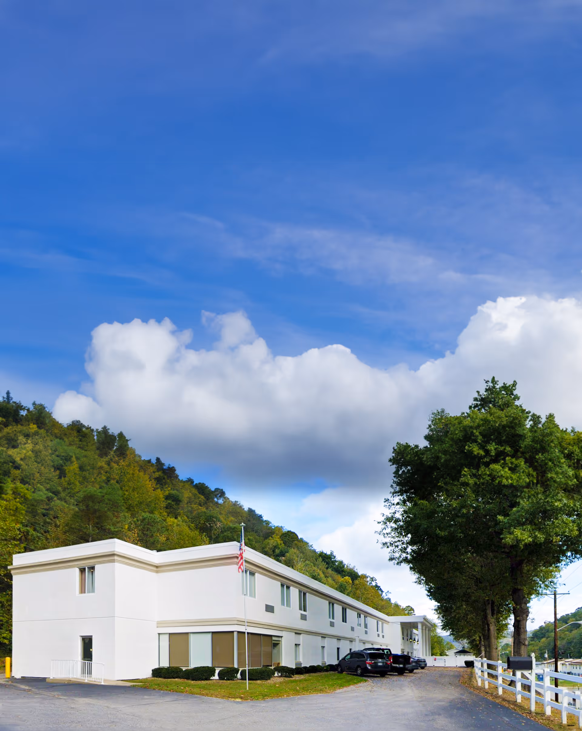 Two-story white senior living building with an American flag, parked cars, trees, and a blue sky with clouds.
