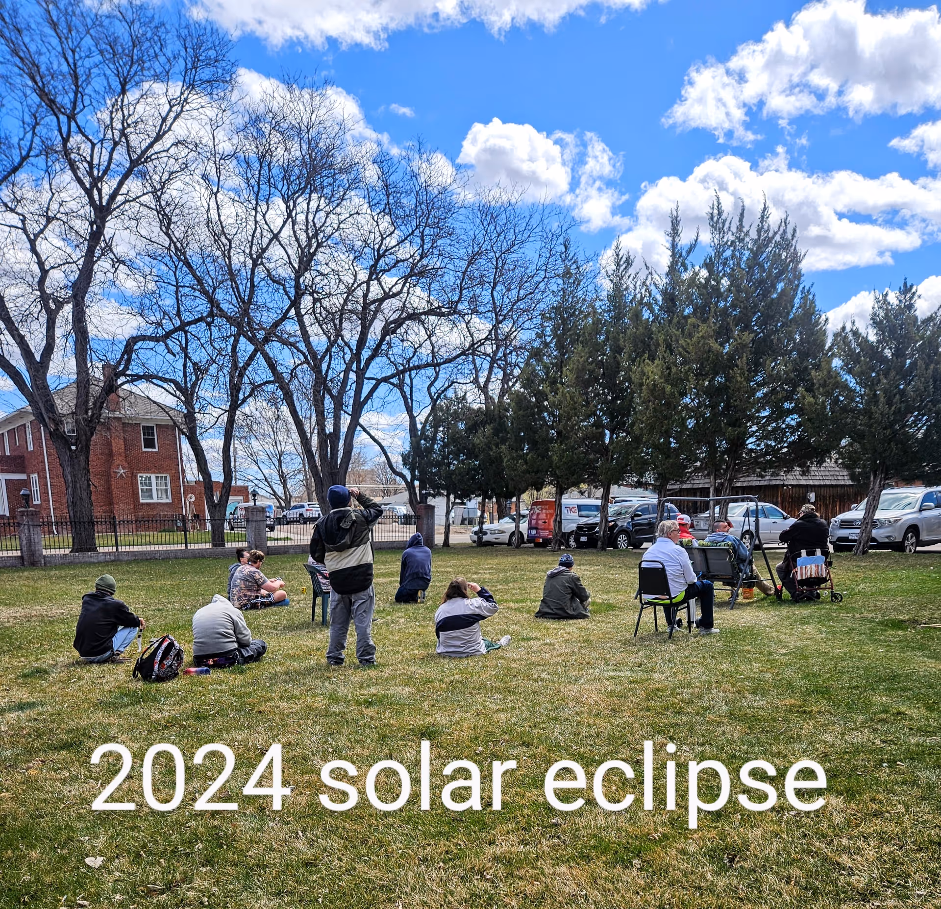 A group of people sitting and standing on a grassy area outdoors, looking up at the sky during a solar eclipse event. The sky is partly cloudy with blue patches visible. Trees and parked cars are in the background.