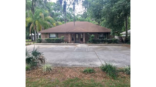 Single-story brick building with a brown roof, shrubbery, and a paved parking lot in front.