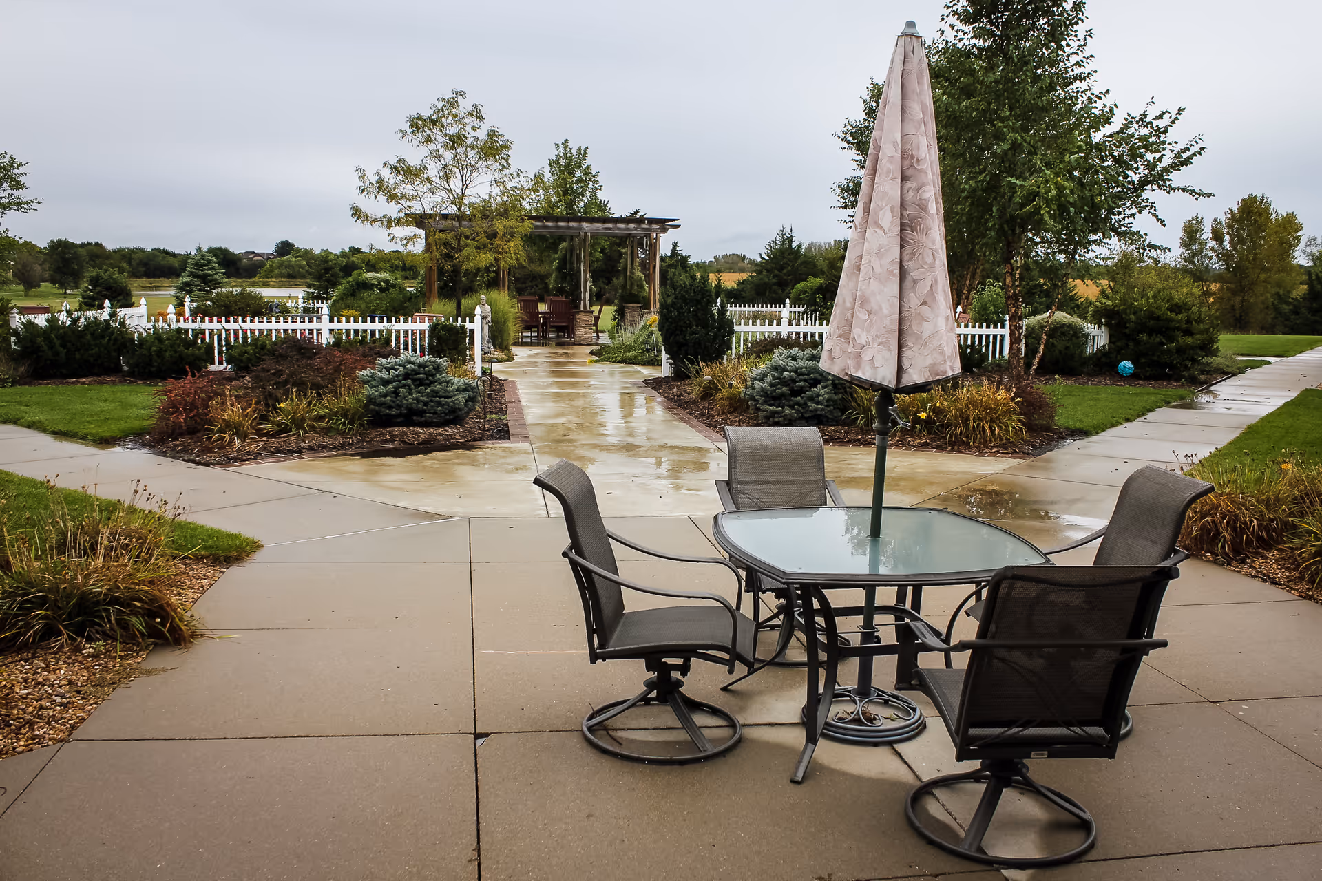 Outdoor patio area with a glass-top table, four chairs, and a closed umbrella. The patio is surrounded by landscaped garden beds, a white picket fence, and a pergola with seating in the background. The ground appears wet from recent rain, and there are trees and greenery beyond the fence.