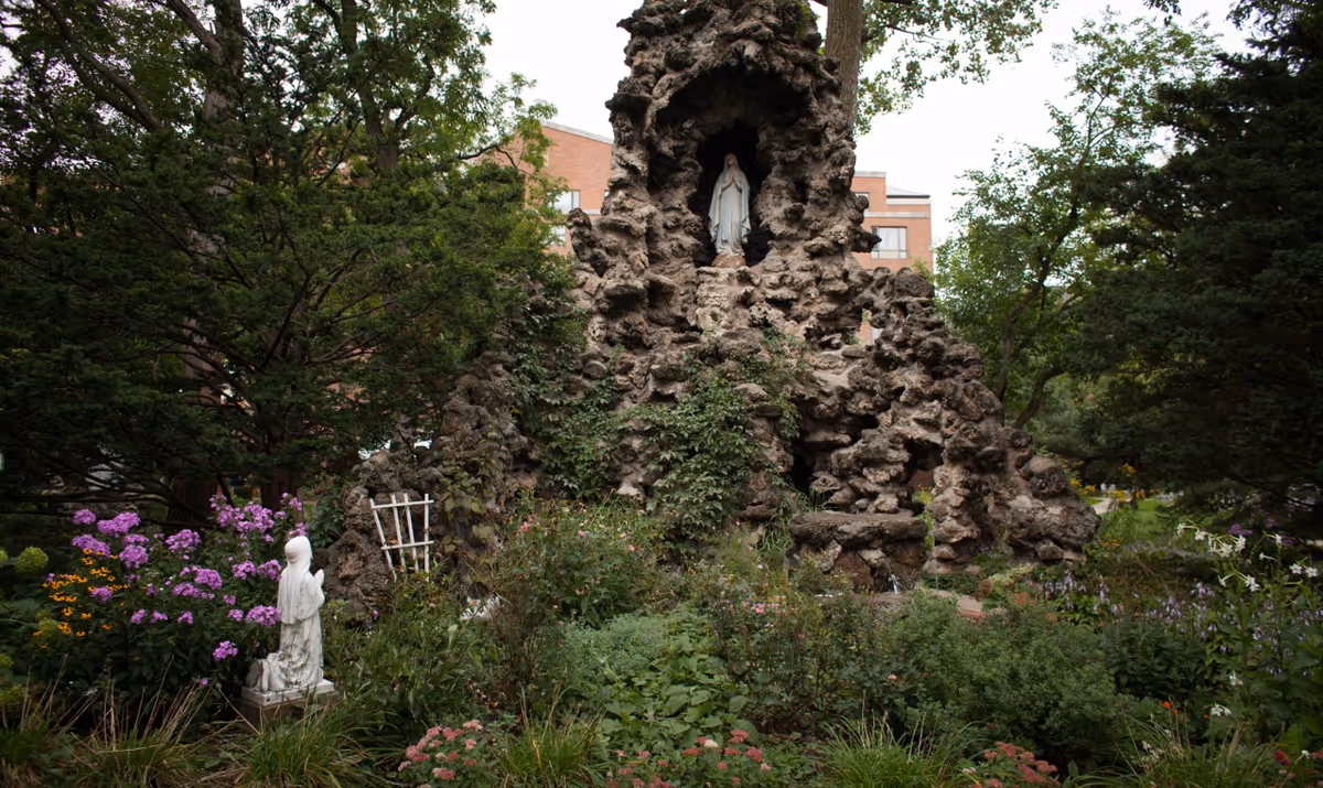 A grotto-like rock formation with a statue of the Virgin Mary surrounded by garden plants and trees in front of a brick building.