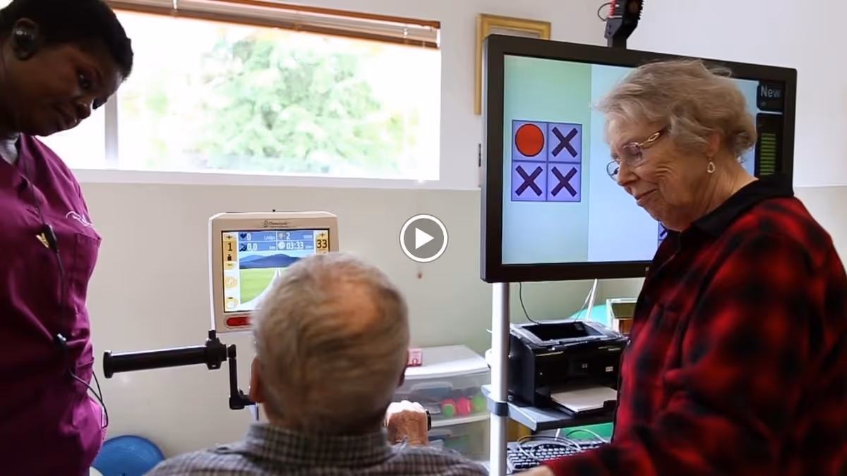 An elderly man and woman interacting with a healthcare worker in a room with medical and computer equipment. The elderly man is seated and looking at a screen, while the elderly woman stands near another screen displaying a tic-tac-toe game. The healthcare worker is standing nearby, observing.
