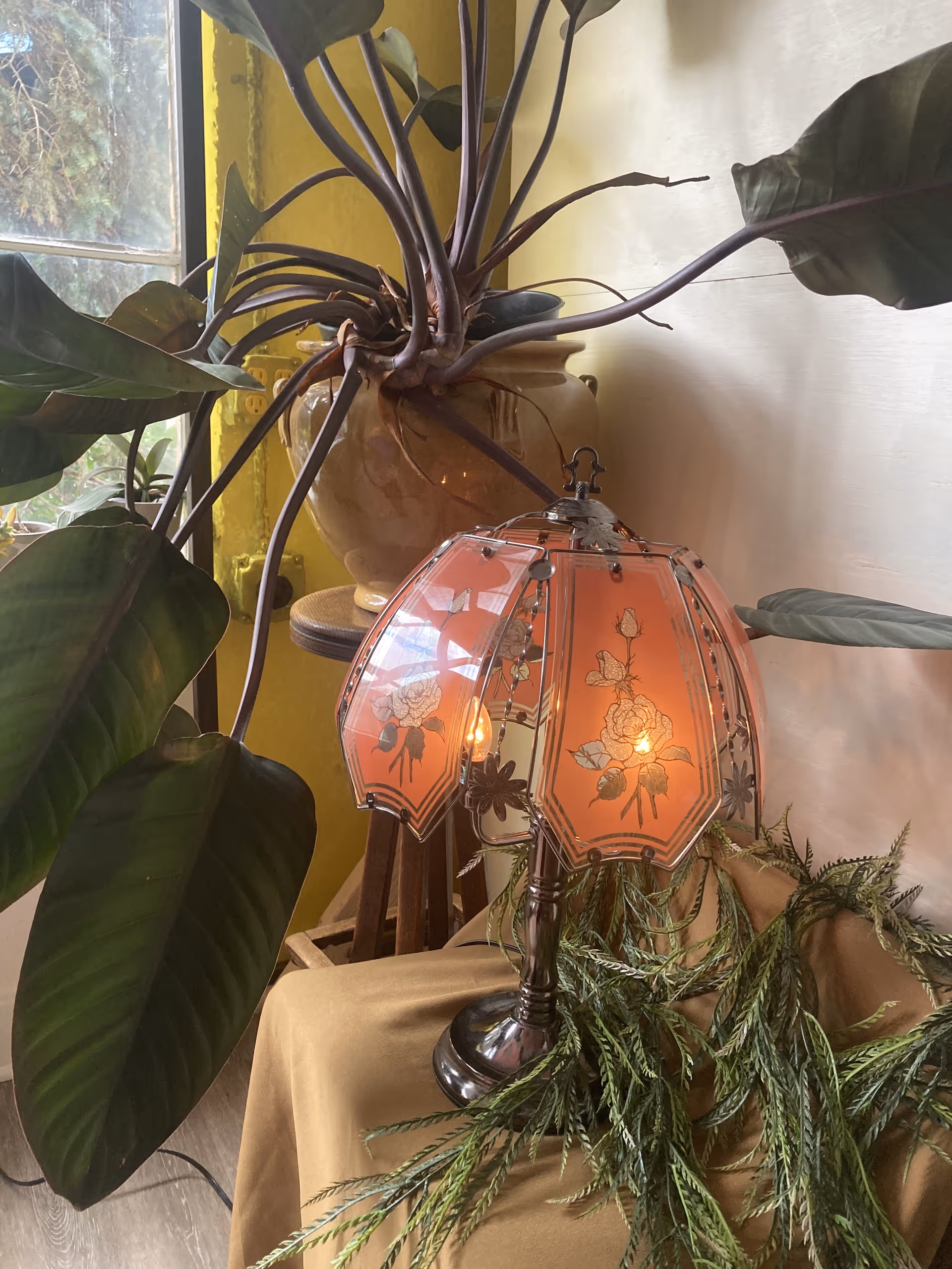 Pink glass lamp beside a large leafy houseplant and beige vase on a cloth-covered table