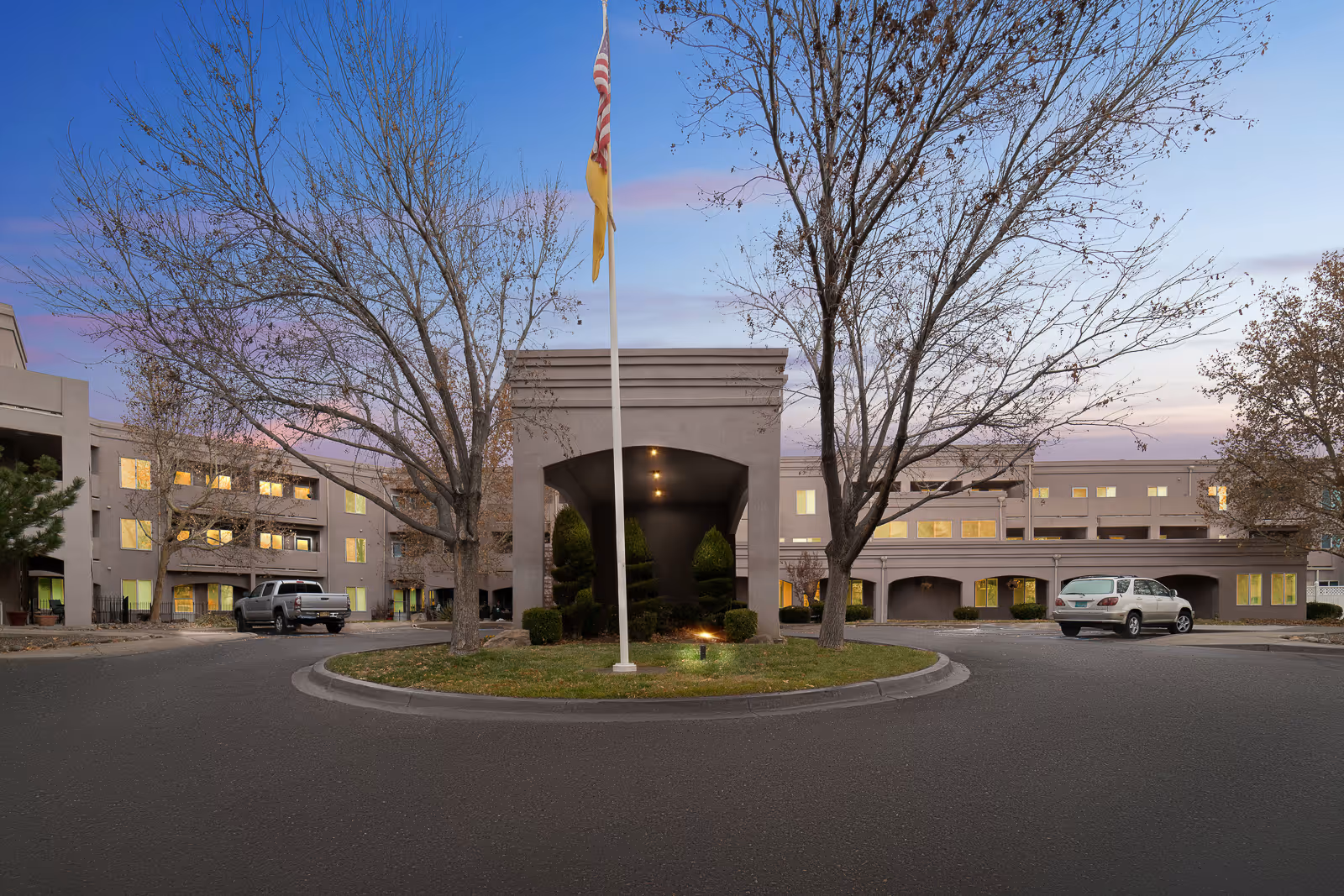 Exterior view of Bear Canyon Estates senior living facility at dusk, showing a circular driveway with a flagpole in the center, leafless trees, and a three-story building with illuminated windows and parked cars.