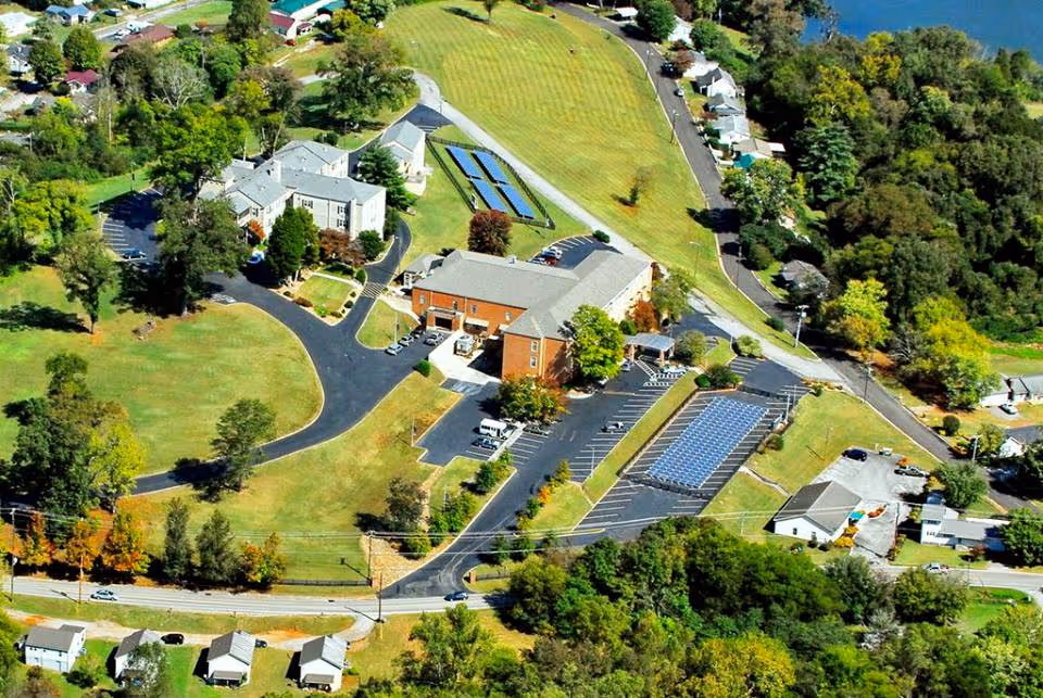 Aerial view of River Oaks Place - Loudon facility showing multiple buildings, parking lots with solar panels, surrounding green lawns, trees, and nearby residential houses.