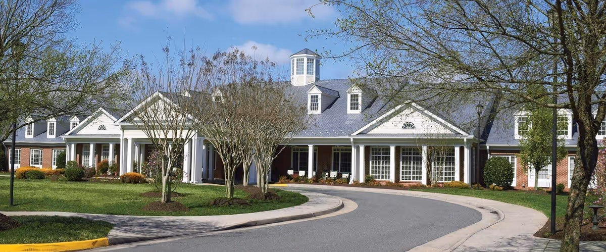 Front entrance of a one-story brick senior living building with white columns, a circular drive, and landscaped grounds.