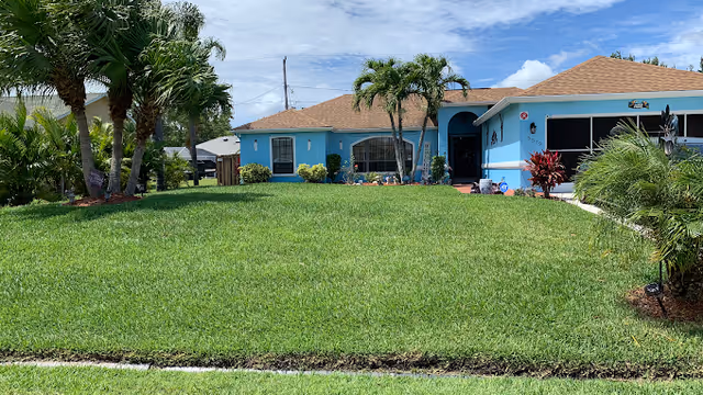Front exterior view of a single-story blue house with a brown shingle roof, surrounded by a well-maintained green lawn and palm trees under a partly cloudy sky.