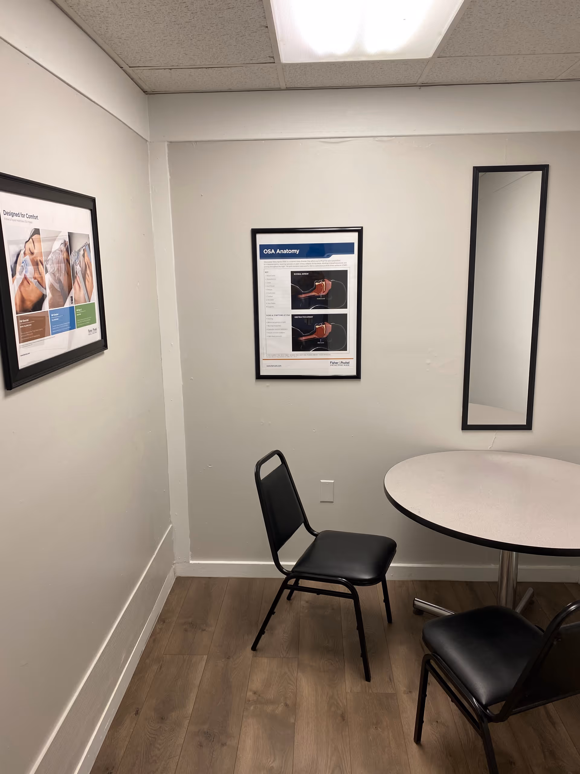 Small interior room with round table, two black chairs, posters, and a tall mirror.