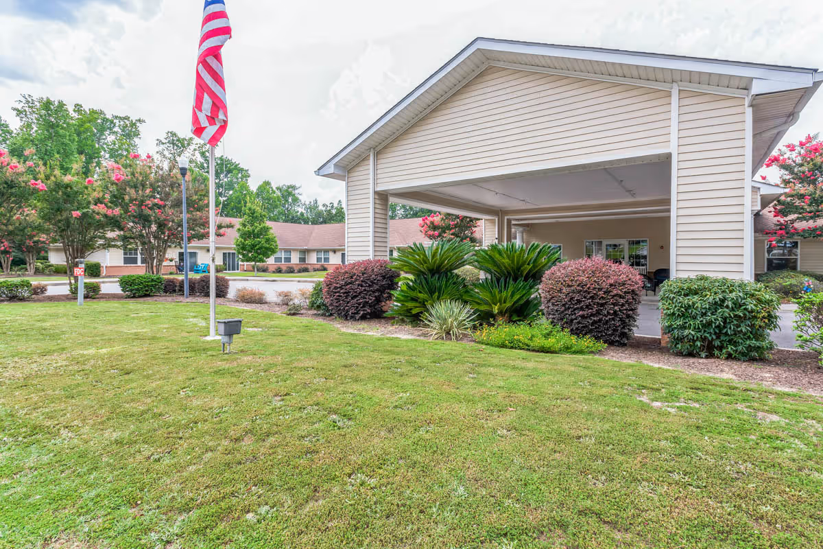Front entrance of a single-story senior living facility with a covered porte-cochere, landscaped lawn, and an American flag.