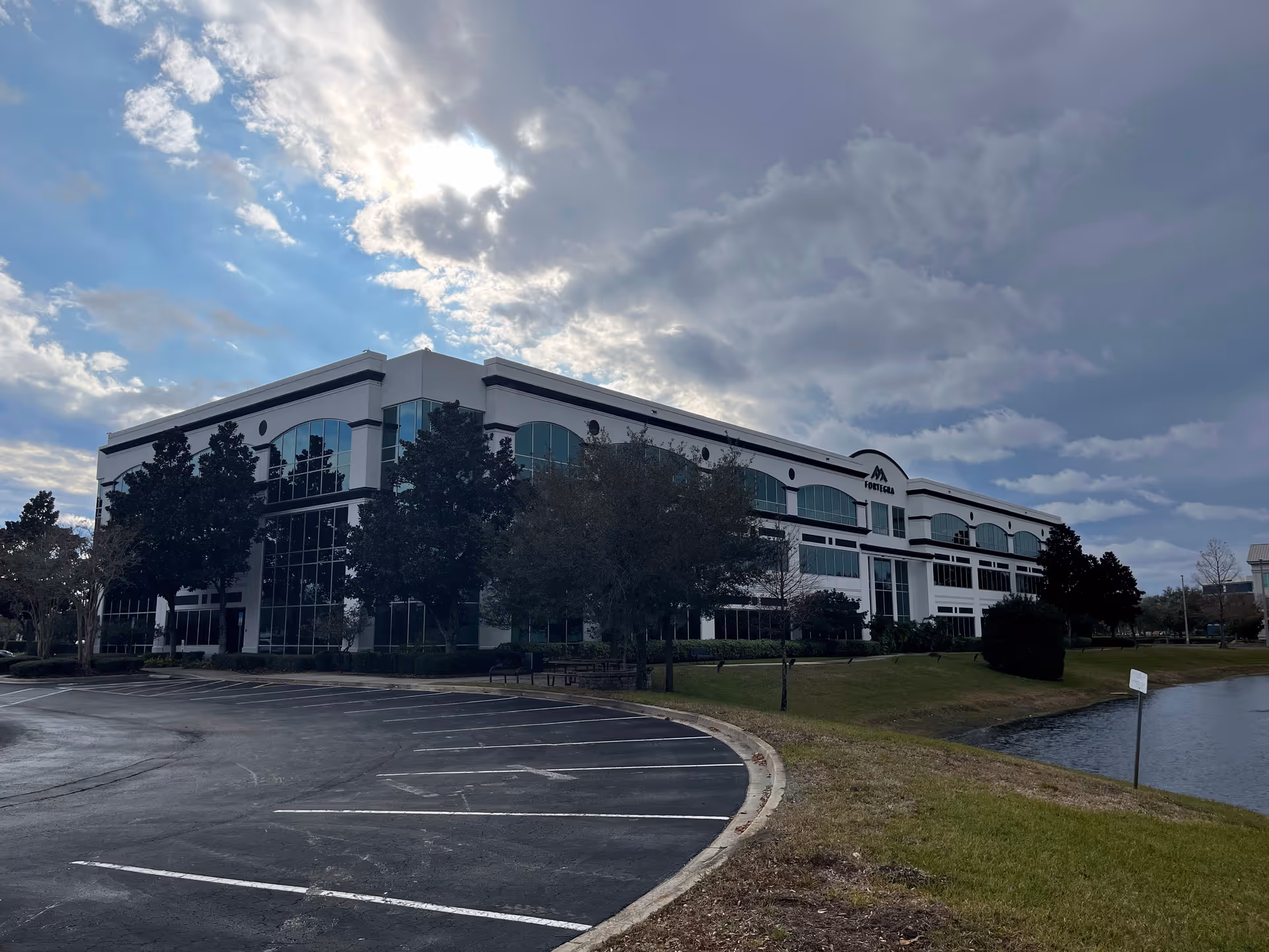 Modern senior living facility with glass-front facade, parking lot, trees, and a pond.