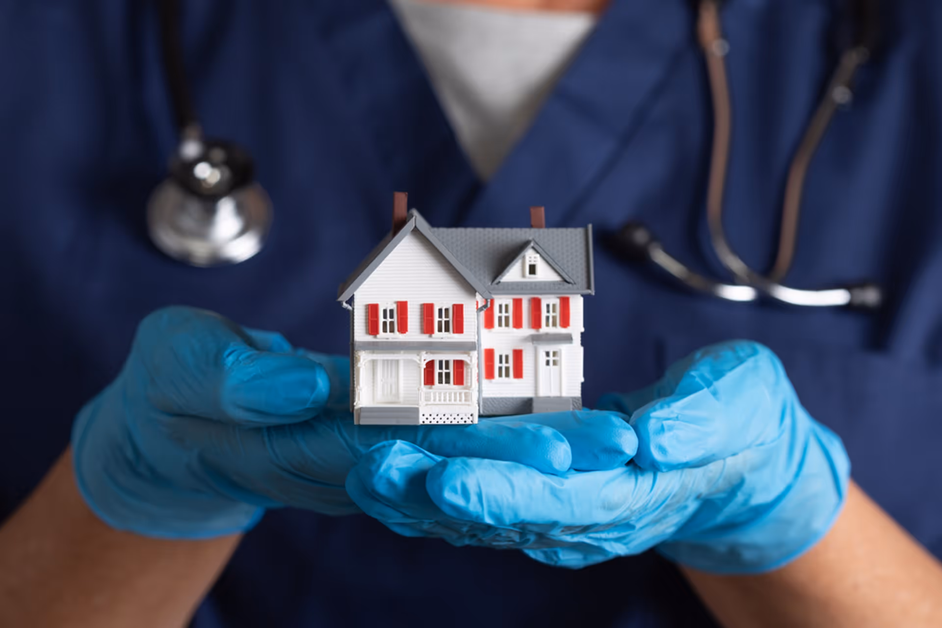 Medical professional in blue gloves holds a miniature house model representing home health care.
