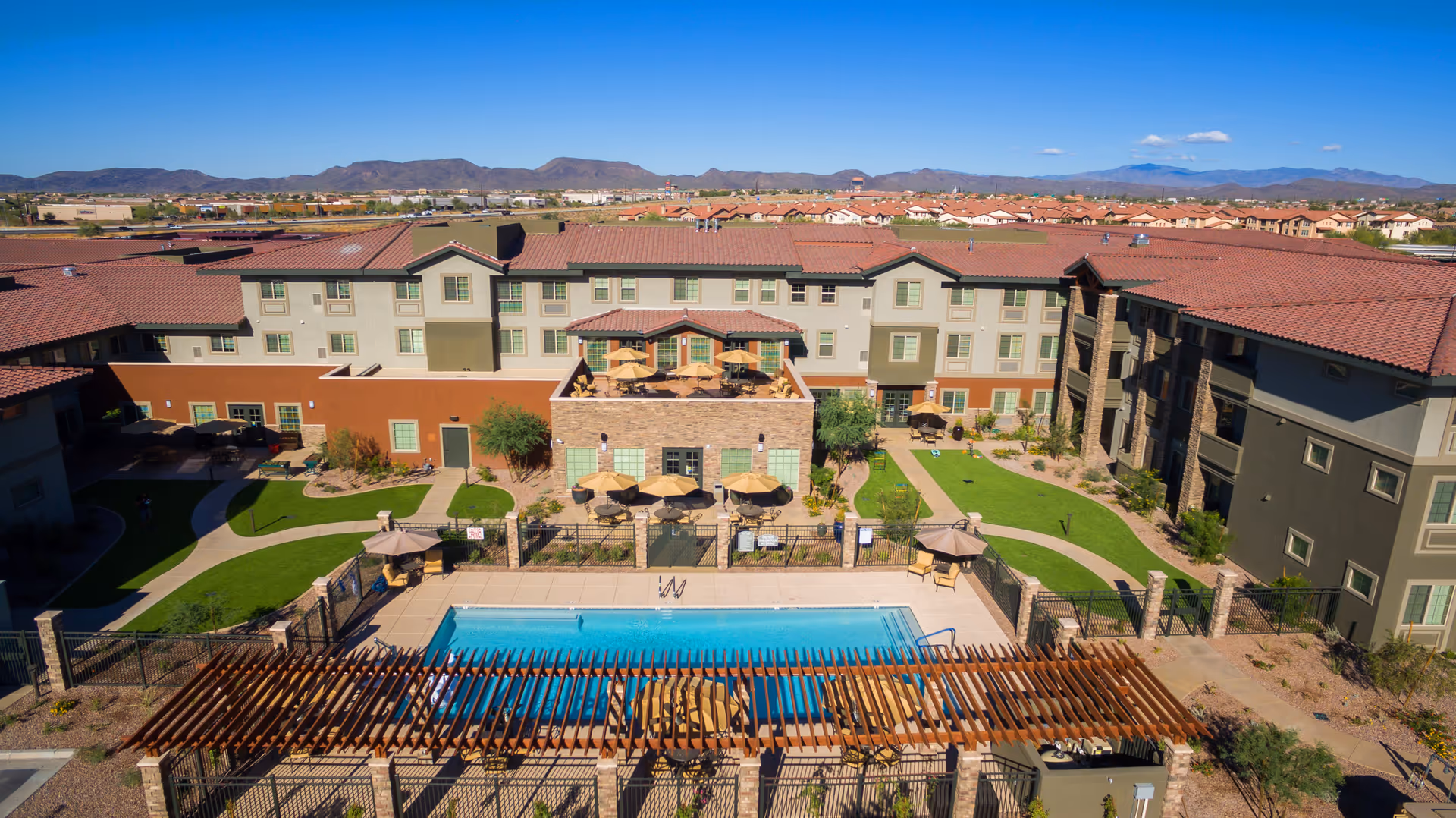 Aerial view of The Enclave at Anthem Senior Living facility showing a large outdoor swimming pool surrounded by lounge chairs and umbrellas. The pool area is fenced and has a wooden pergola in the foreground. The multi-story building with red-tiled roofs and green-trimmed windows surrounds the pool area. There are landscaped green lawns and walking paths around the building. Mountains and a clear blue sky are visible in the background.