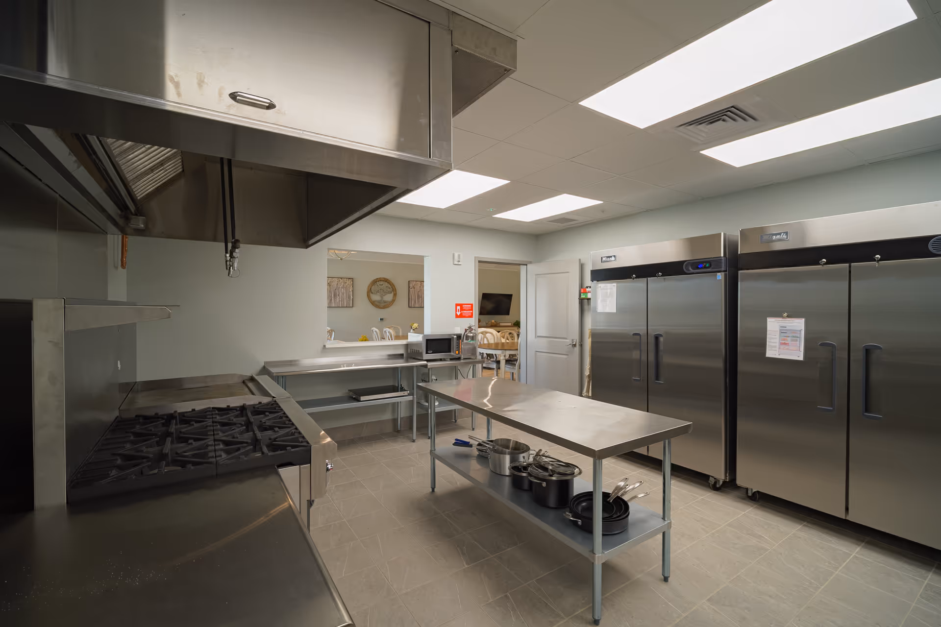 Industrial stainless steel kitchen with large appliances, prep island, and overhead ventilation in a senior facility.