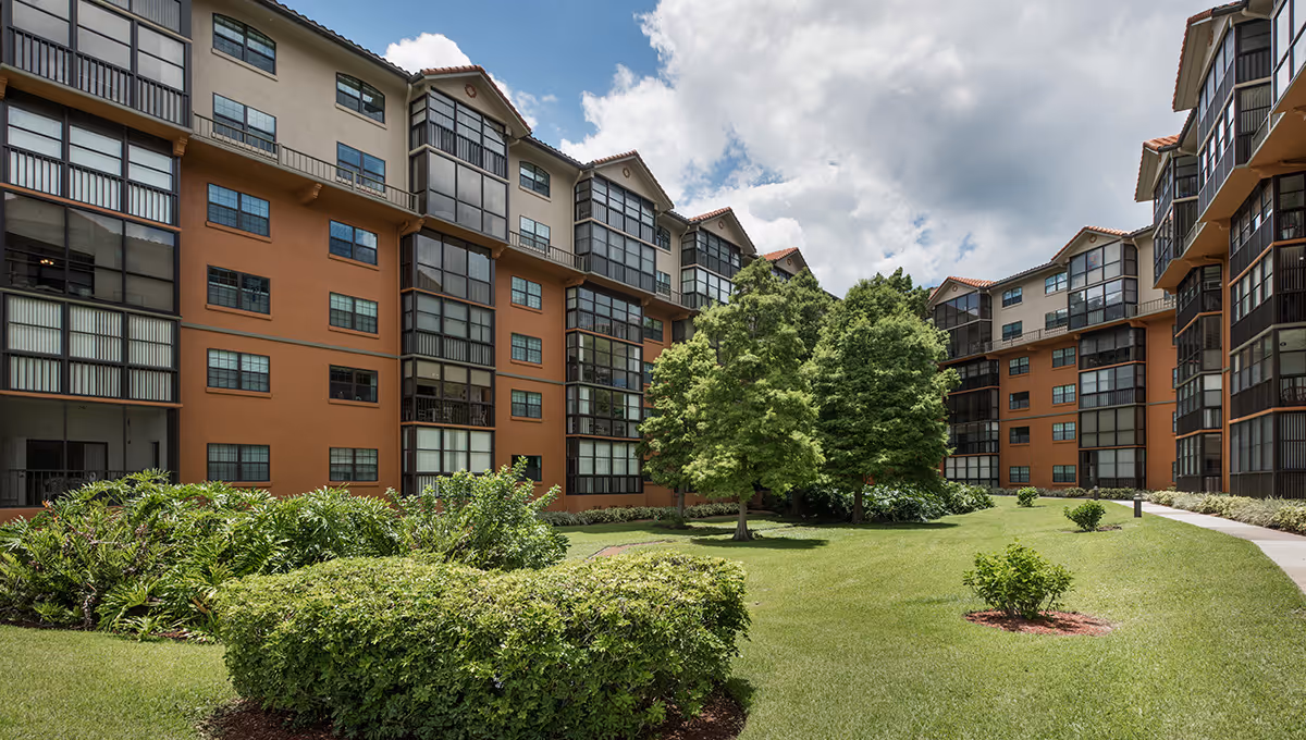 Multi-story senior living building surrounding a landscaped grassy courtyard with trees and shrubs under a partly cloudy sky.