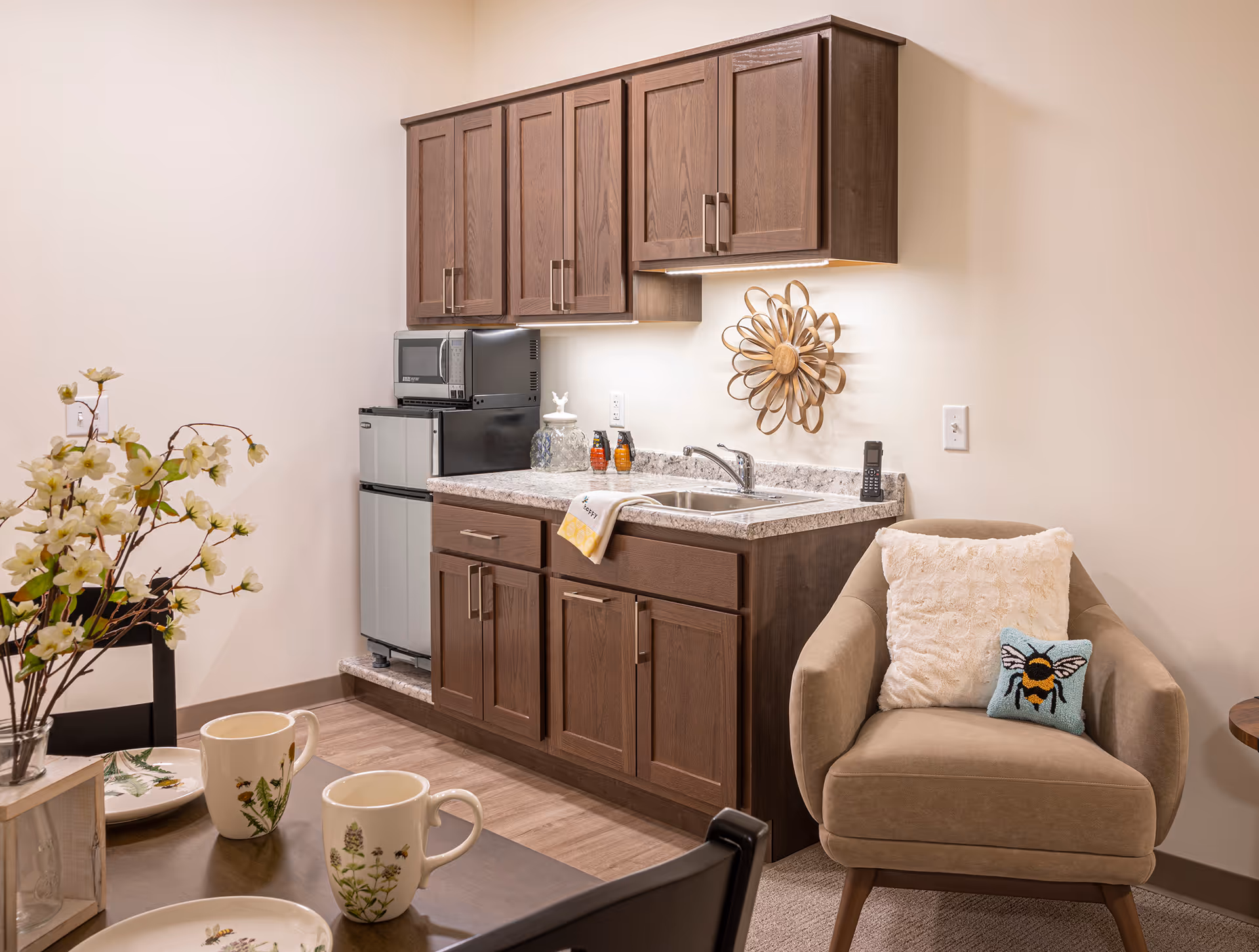 Compact kitchenette with dark cabinets, sink, mini-fridge, microwave, and a beige armchair nearby.