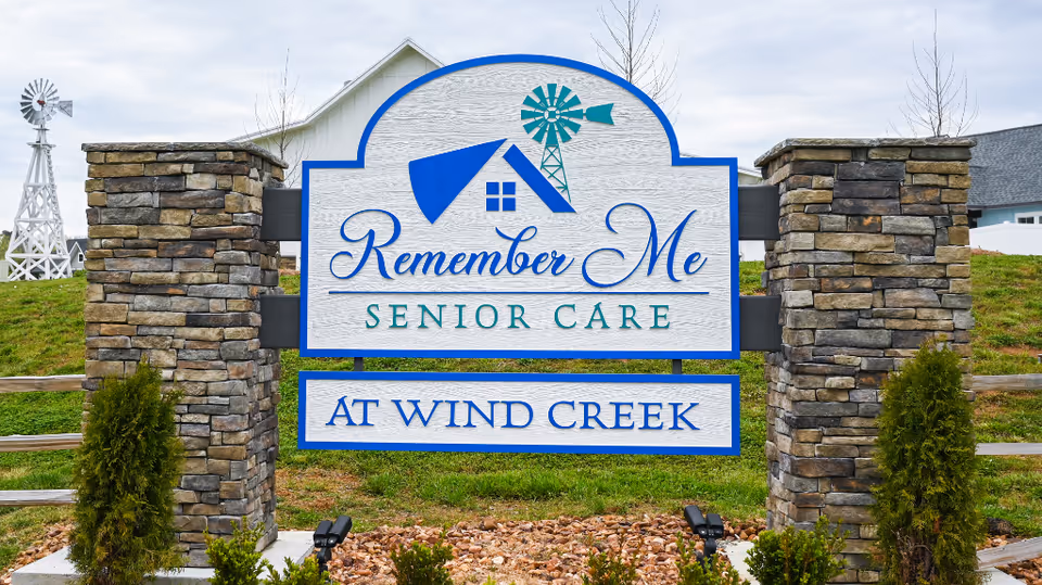 A large outdoor sign for Remember Me Senior Care at Wind Creek, mounted between two stone pillars with a grassy area and a white windmill in the background.
