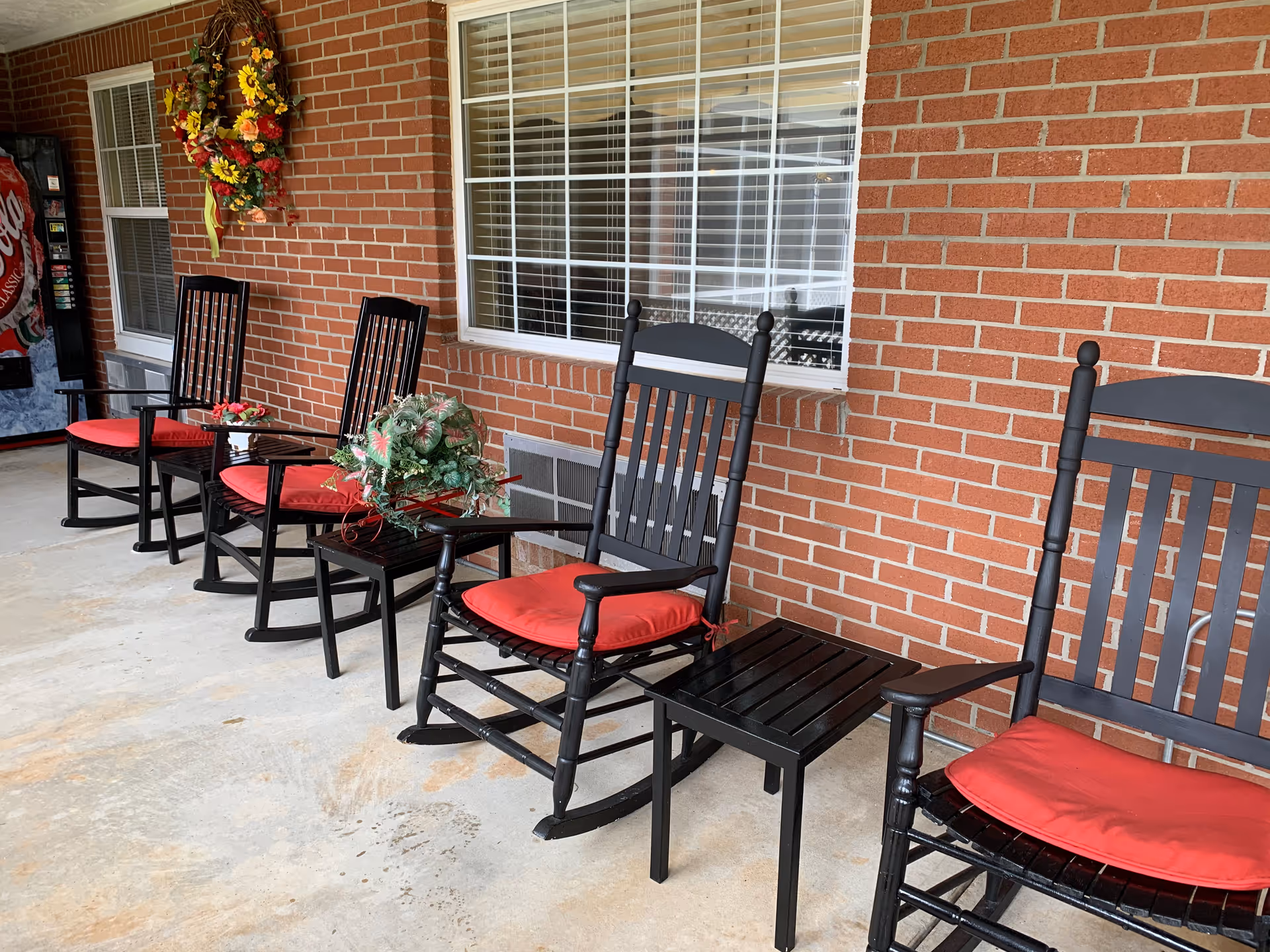 A row of black wooden rocking chairs with red cushions lined up against a brick wall under a covered porch. There are small black tables between some of the chairs, decorated with floral arrangements. A window with white blinds and a decorative wreath with sunflowers hang on the brick wall. A vending machine is visible in the background.