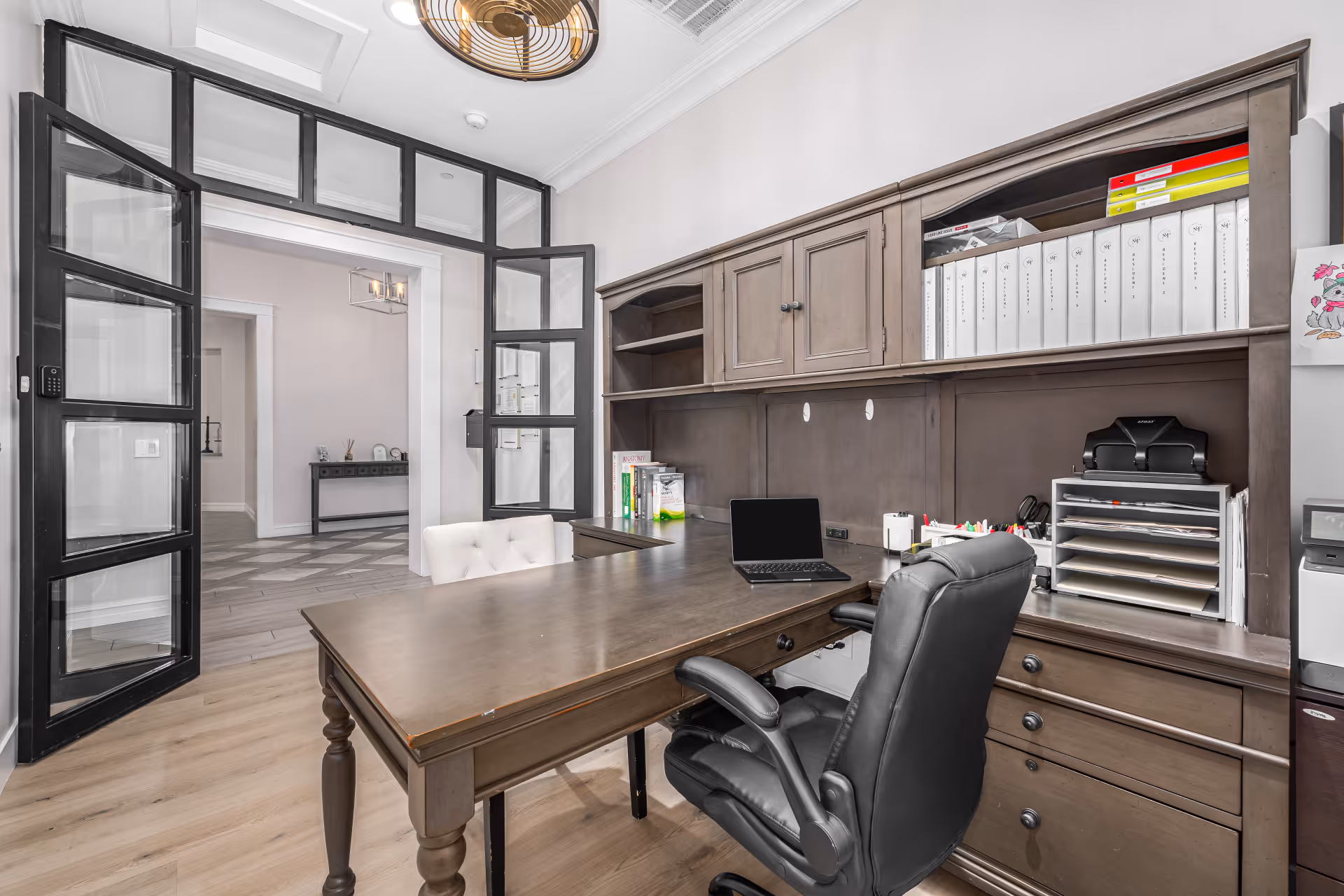A modern office space with a large wooden desk, a black leather office chair, and a white cushioned chair. The desk has a laptop, office supplies, and organized binders on shelves above. The room features glass-paneled double doors leading to a hallway with light-colored walls and flooring.