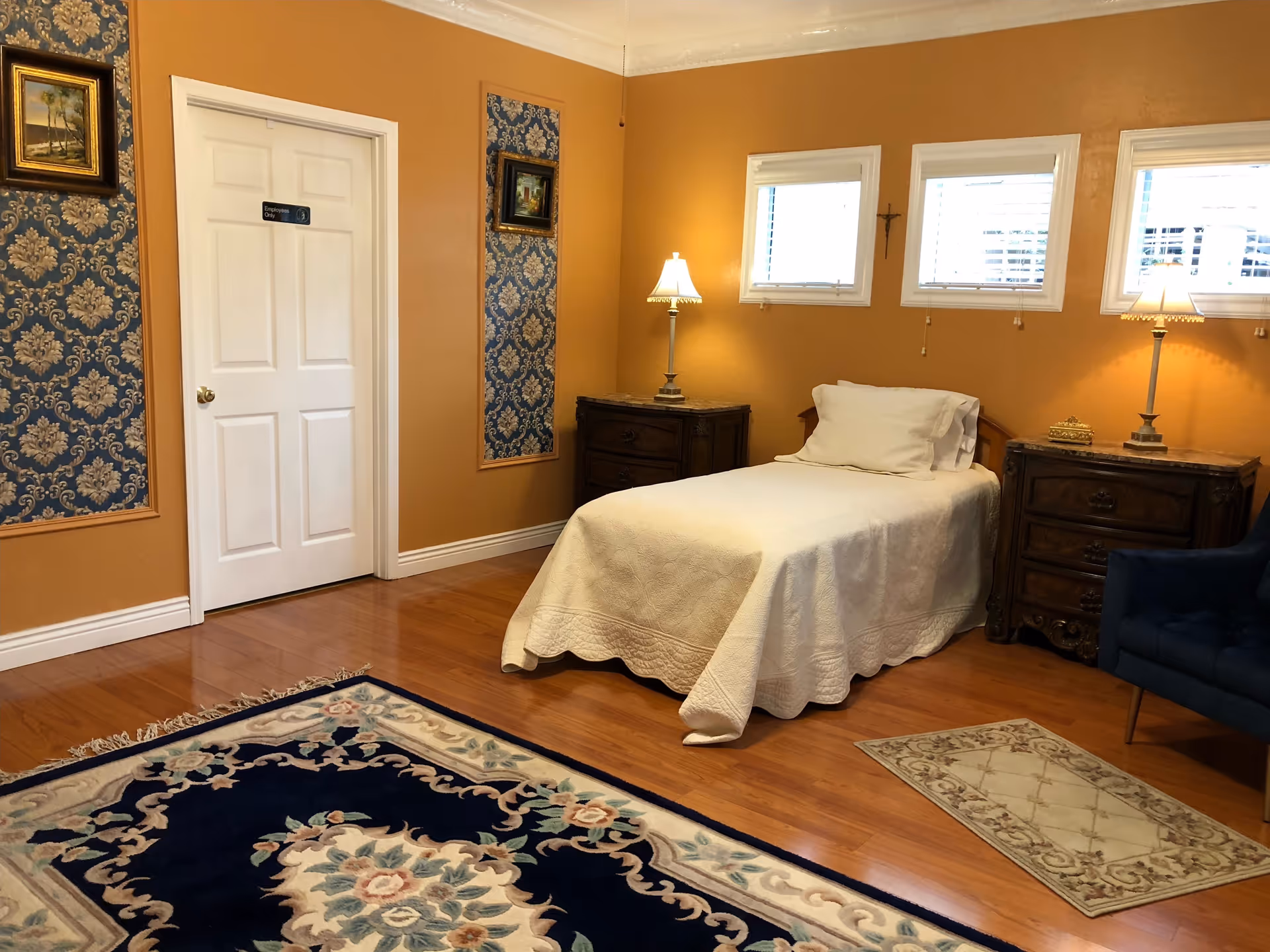 A cozy bedroom with a single bed covered in a white quilt, two wooden nightstands with lamps on either side of the bed, three small windows with blinds above the bed, a blue armchair, and two framed paintings on the orange walls with blue patterned wallpaper panels. The floor is wooden with two decorative rugs.