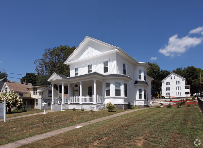 White two-story house with wraparound porch on a grassy lot, clear blue sky.