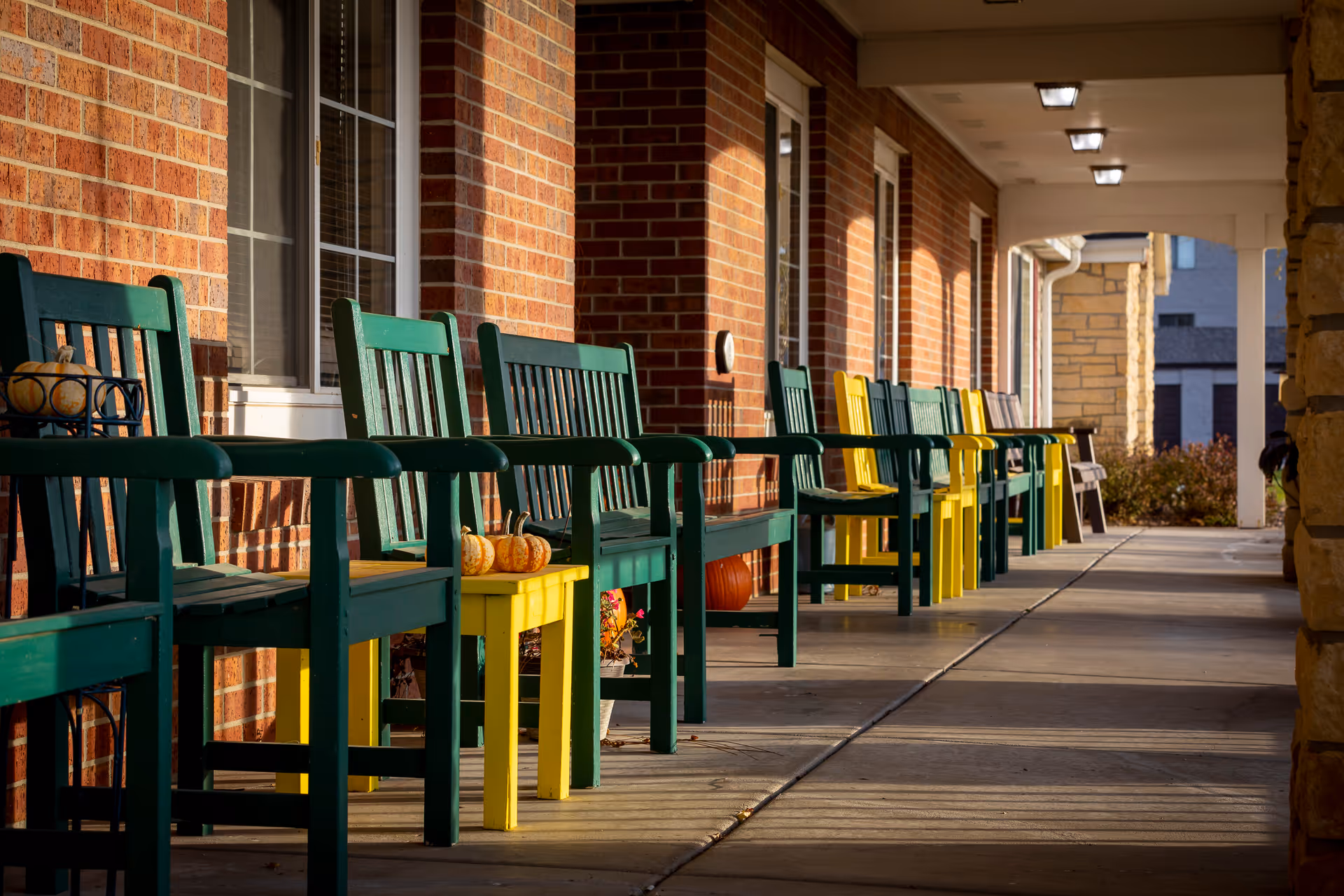 A covered outdoor walkway at Homestead Assisted Living of Hastings with a row of green and yellow wooden chairs and small tables placed against a brick wall. Small decorative pumpkins are placed on some tables, and the area is lit by warm sunlight.