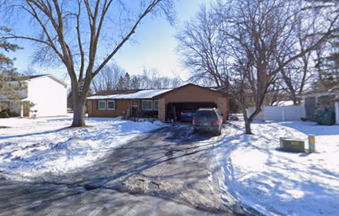 Snow-covered front of a single-story house with a driveway and a parked car.
