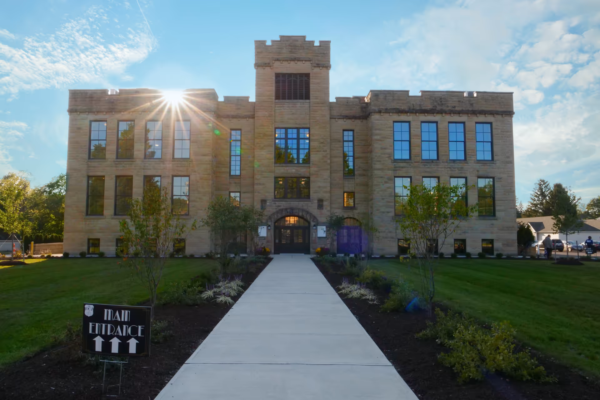 Front exterior of a large stone three-story school-like building with a walkway leading to the main entrance and sun glare in the sky.