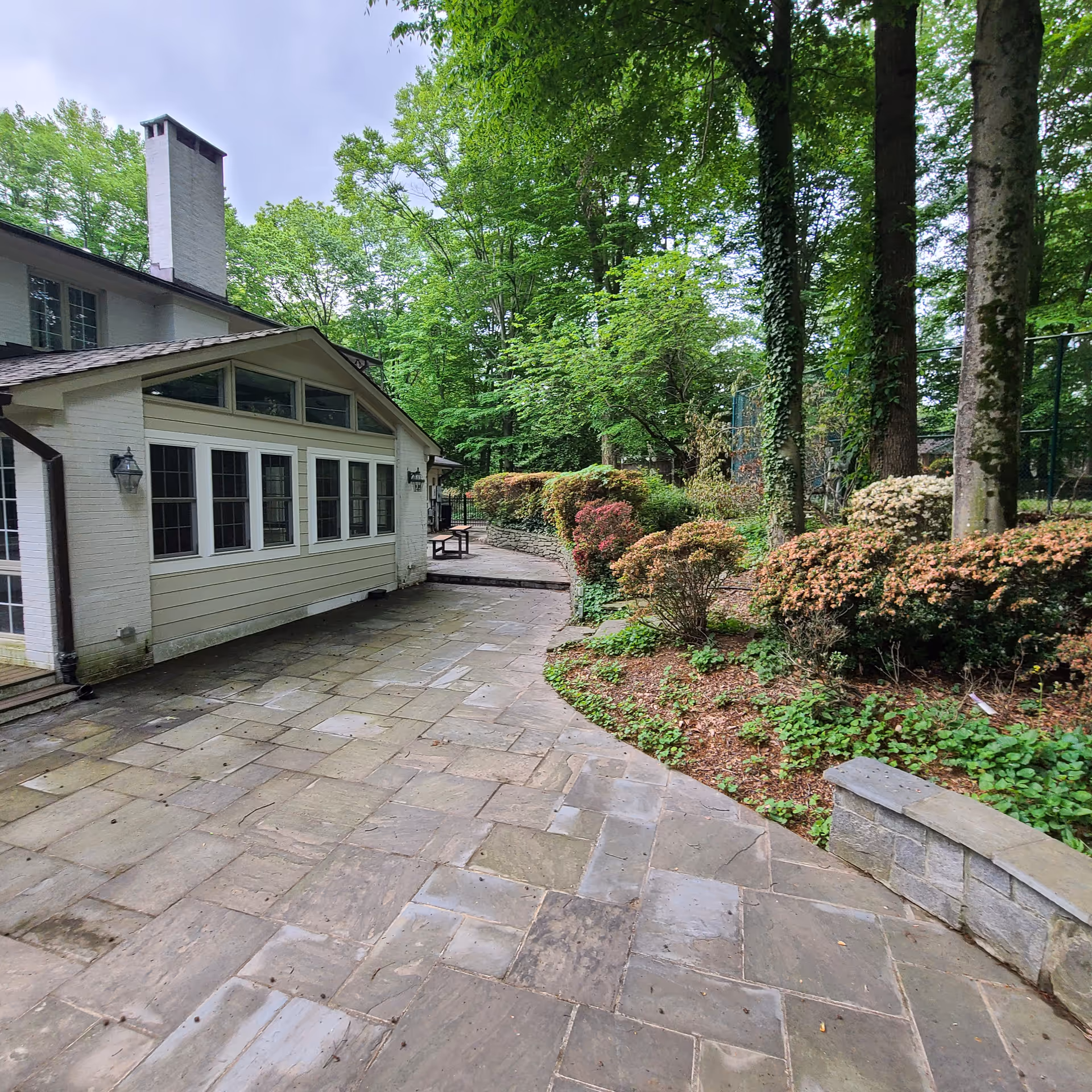 Stone patio beside a white building, bordered by lush trees and flowering shrubs.