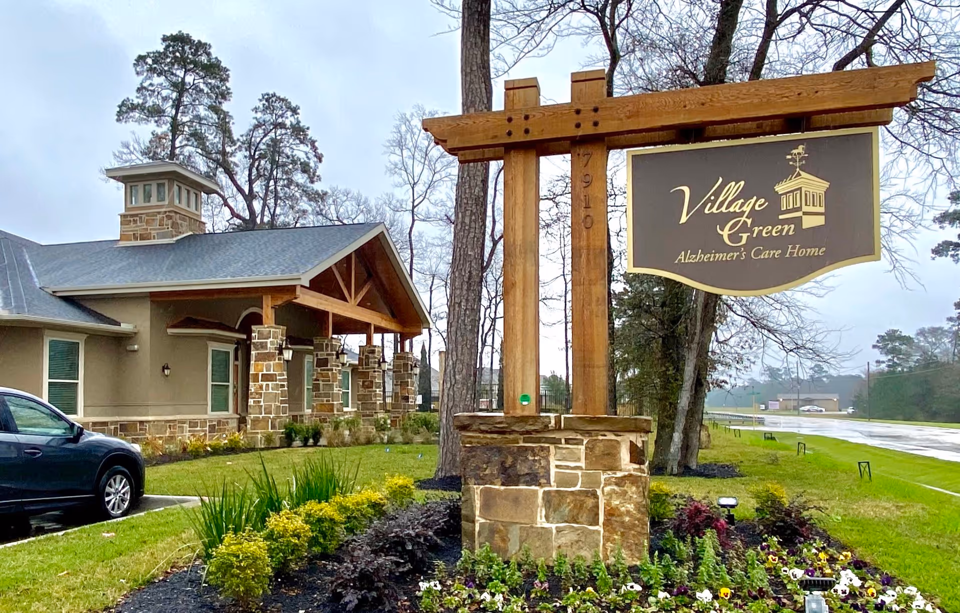 Exterior view of Village Green Alzheimer's Care Home building with a stone and wood signpost in front displaying the facility name. The building has a stone and beige facade with a covered porch and a parking area with a black vehicle. The surrounding area includes landscaped greenery and trees under a cloudy sky.