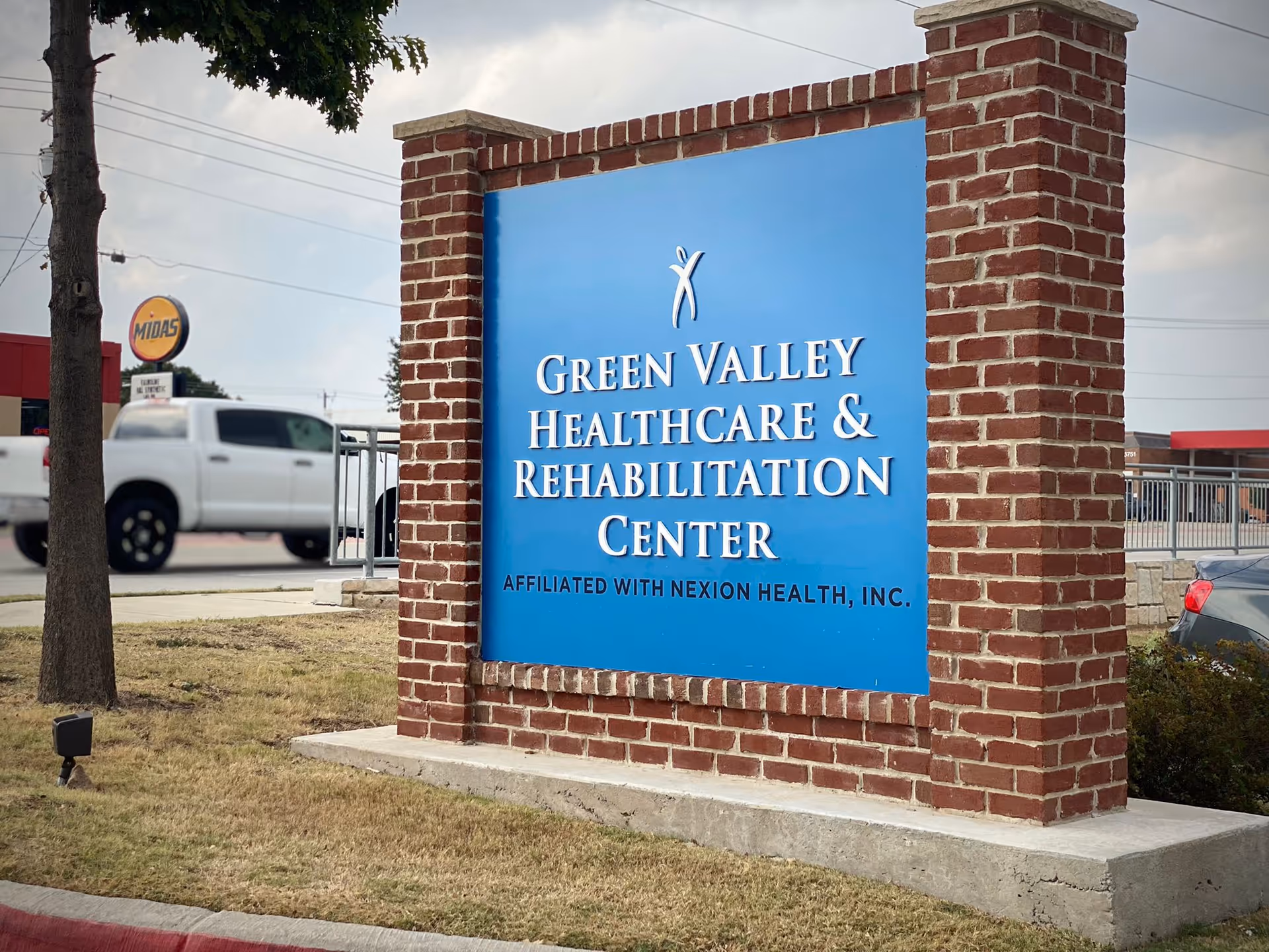 Brick-framed blue sign reading Green Valley Healthcare & Rehabilitation Center beside a street.