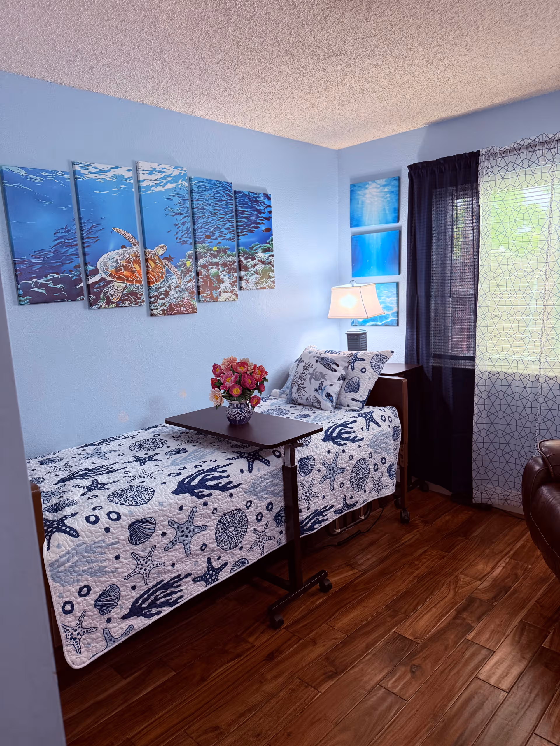 Blue-themed bedroom with a single bed, sea-print quilt, lamp, flowers, and wall art.