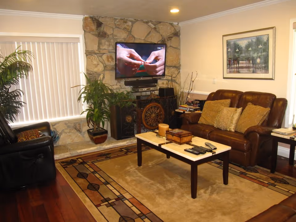 A cozy living room with a stone fireplace wall featuring a mounted flat-screen TV. The room has a brown leather sofa with multiple cushions, a black leather recliner, a coffee table with candles, remote controls, and a decorative box. There are two potted plants near the window with vertical blinds, a framed painting on the wall, and a patterned area rug on the wooden floor.