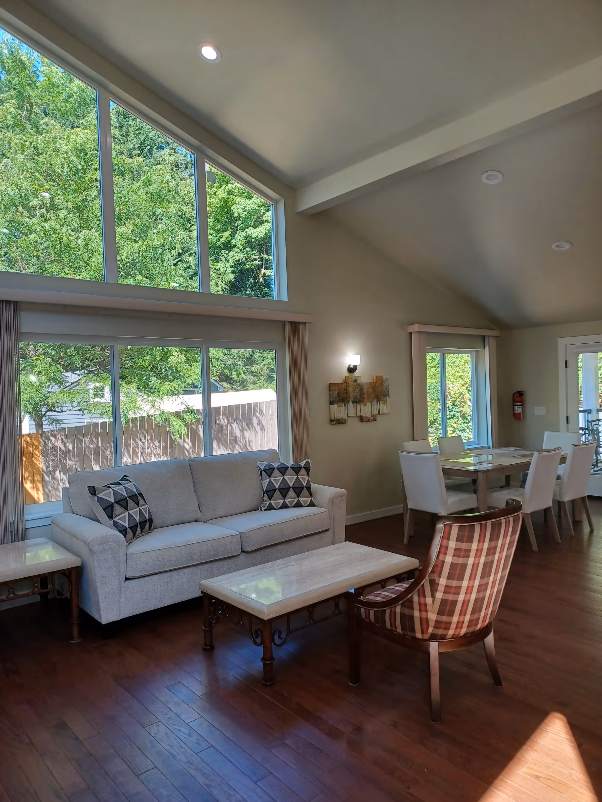Bright open living and dining area with large windows, a light sofa, coffee table, plaid armchair, and wood floors.