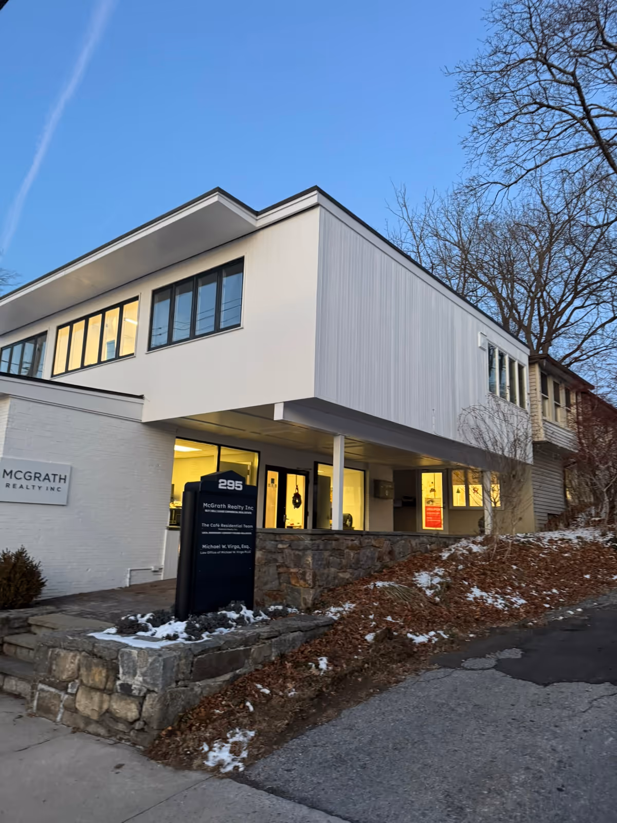 Modern two-story building with large windows, white facade, and a stone retaining wall along the street.