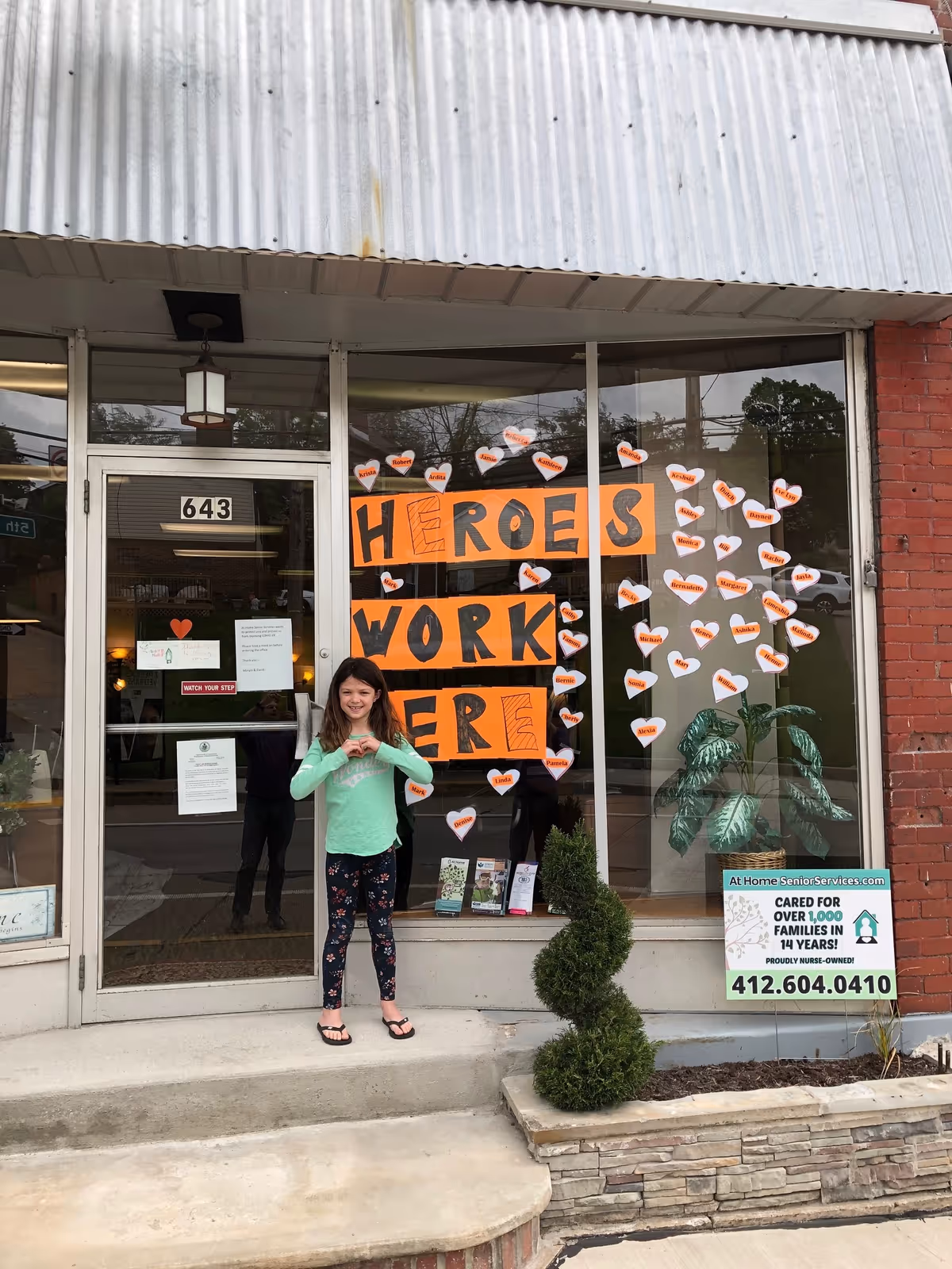 Storefront of At Home Senior Services with 'Heroes Work Here' sign and a girl standing outside.