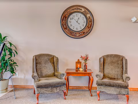 Two upholstered chairs with a small table and decorative clock in a beige lounge.