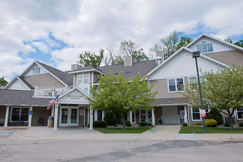 Exterior view of Riverfront Manor senior living facility with entrance, trees, and American flag.
