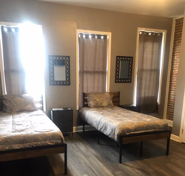 A bedroom with two single beds, patterned bedding, three tall windows with curtains, and a small nightstand between the beds.