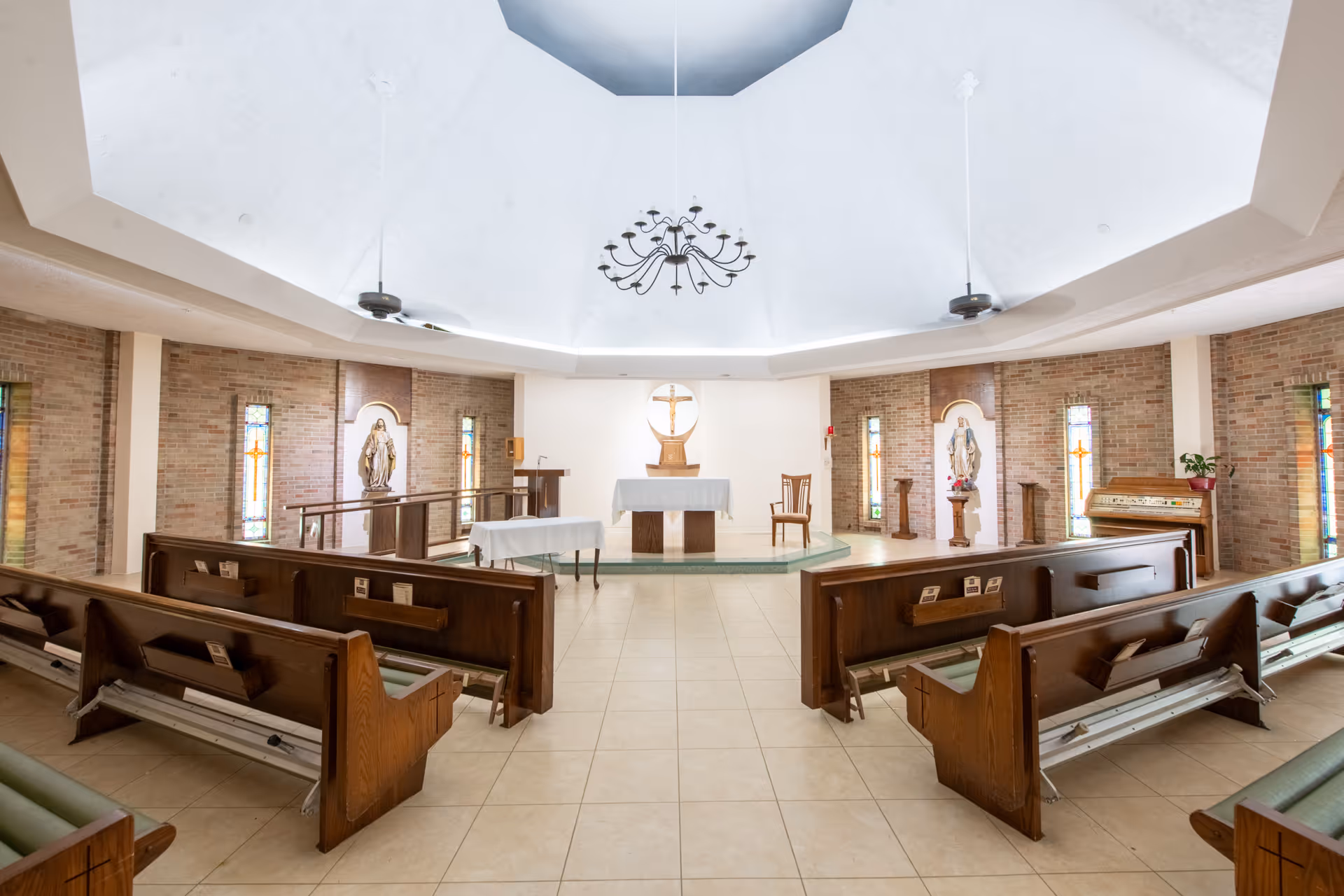 Interior of a chapel with wooden pews facing an altar, stained glass windows, religious statues, and a domed ceiling.