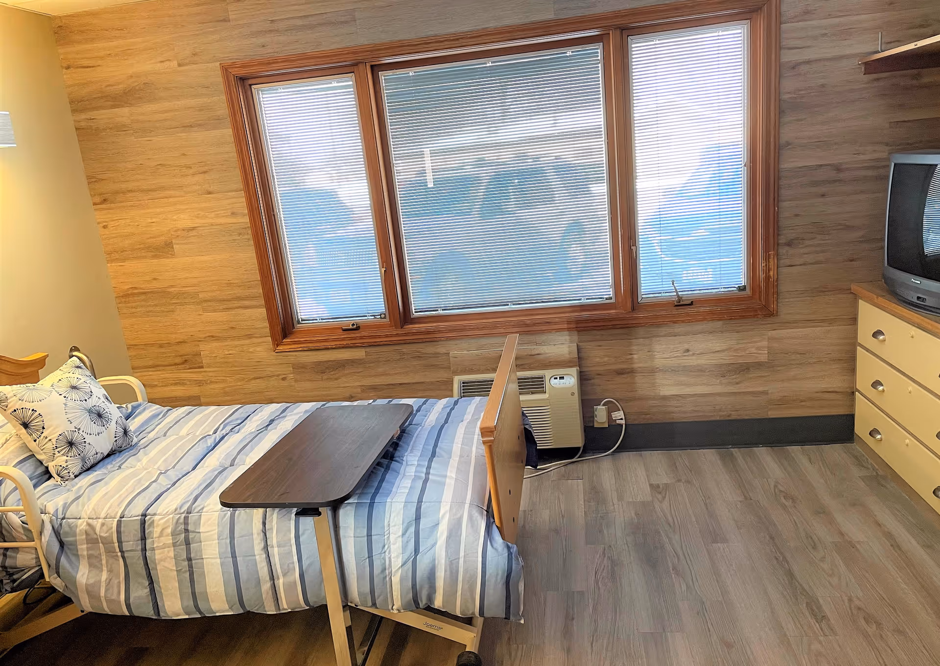 A bedroom in a senior living facility with a single bed covered in a blue and white striped blanket and a floral pillow. A wooden overbed table is positioned on the bed. The room has wood-paneled walls, a large window with blinds, a beige dresser with drawers, and an older-style television on top. The floor is covered with light wood laminate.