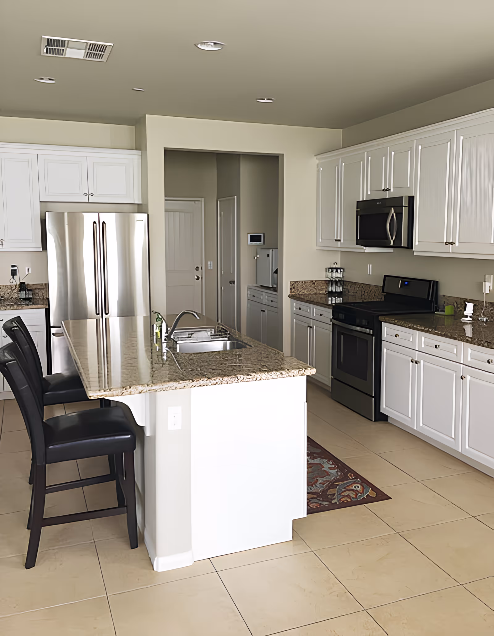 Modern kitchen with white cabinetry, granite countertops, a stainless steel refrigerator, stove, and microwave. There is a kitchen island with a sink and two black chairs.