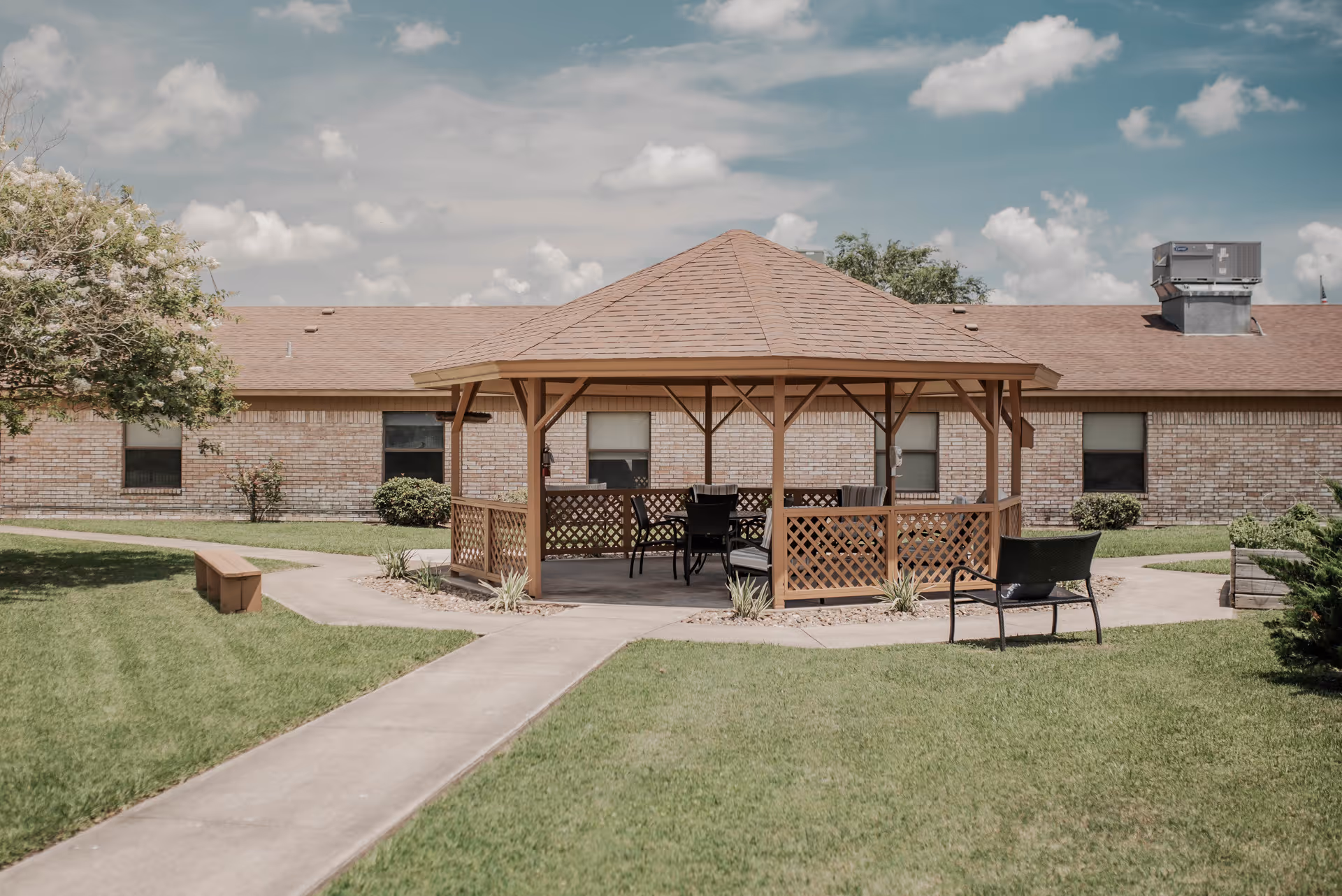 Outdoor gazebo with a wooden roof and lattice sides situated on a concrete path surrounded by green grass. There are chairs and a table inside the gazebo. In the background, there is a single-story brick building with several windows and a clear sky with some clouds.