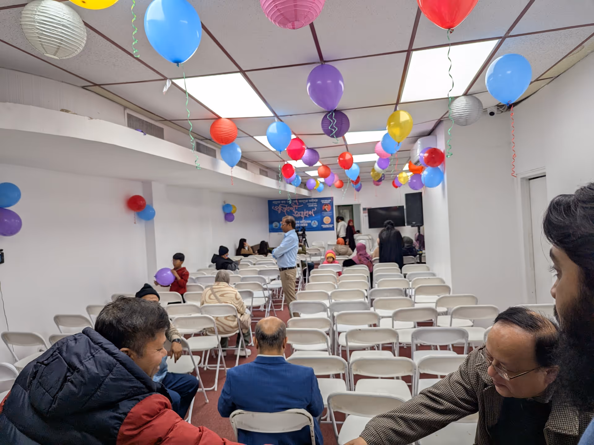 A decorated indoor hall with balloons, rows of white chairs, and attendees.