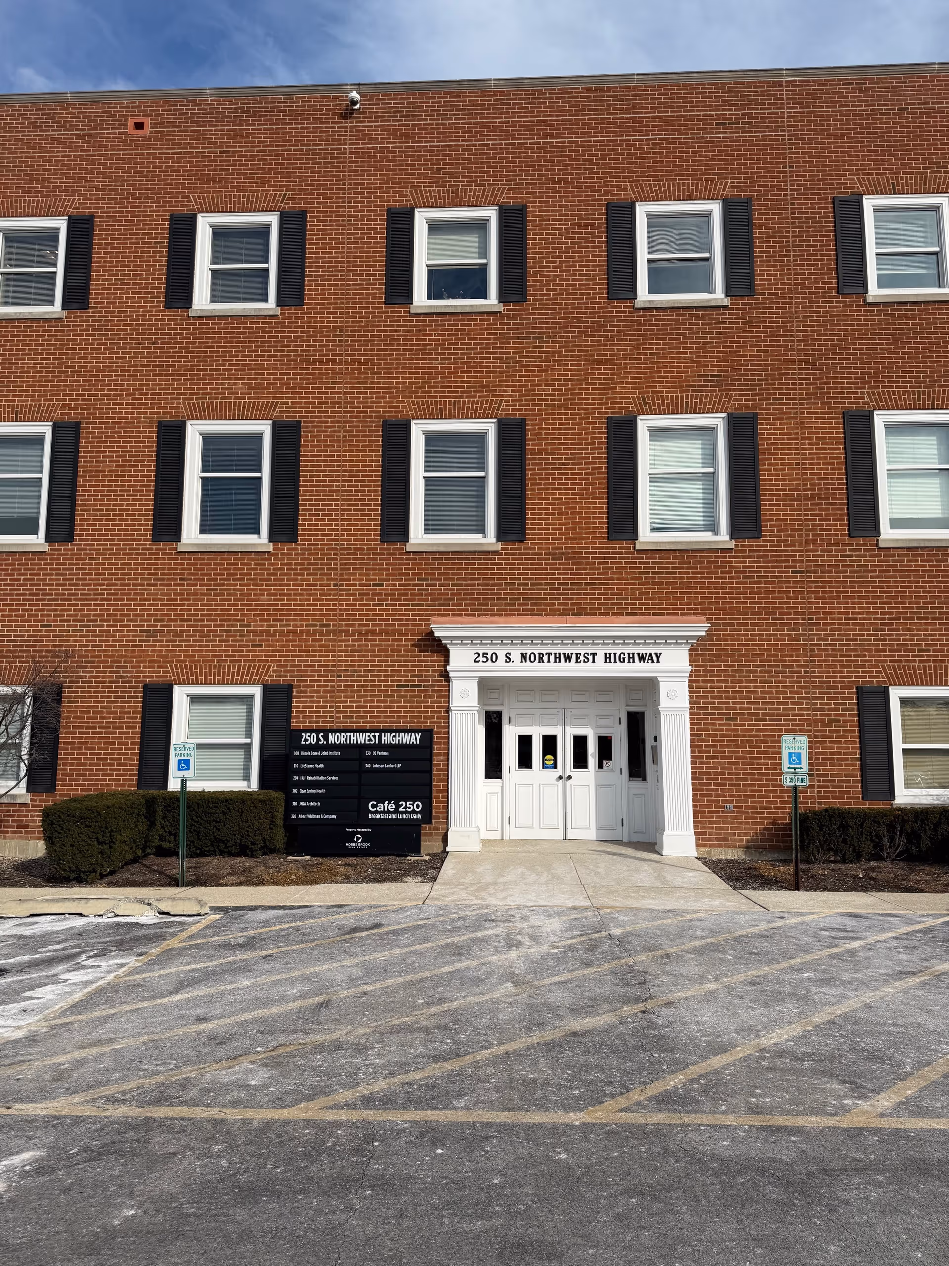 Brick three-story building with white entrance, black shutters, and parking lot in foreground.