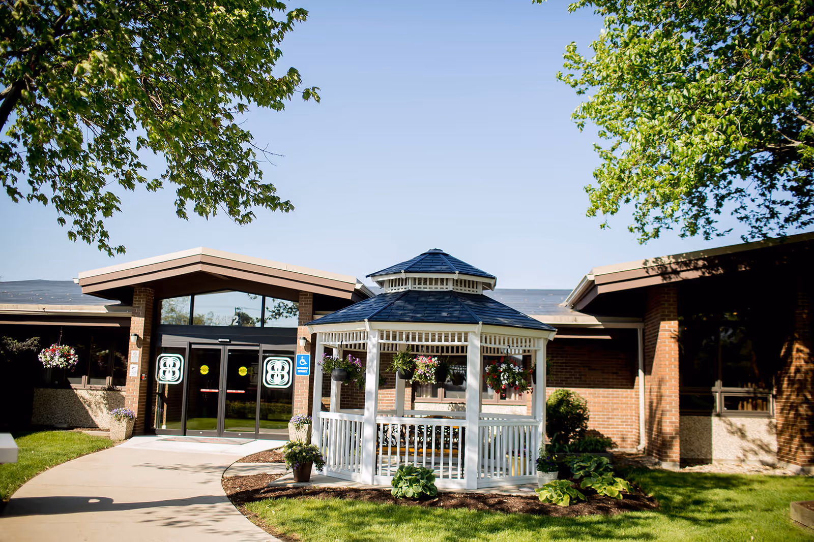 Brick senior living facility entrance with glass doors and a white gazebo surrounded by greenery